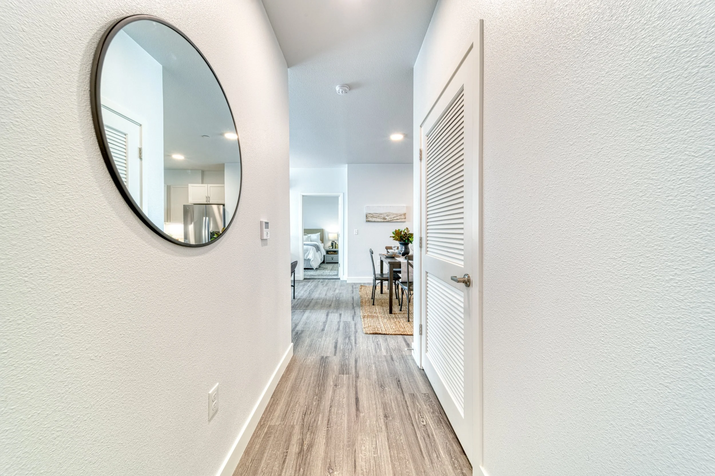 Interior hallway leading to a bedroom with visible bed and nightstand, and a small dining area with a table and chairs, modern decor, wood flooring, white walls.