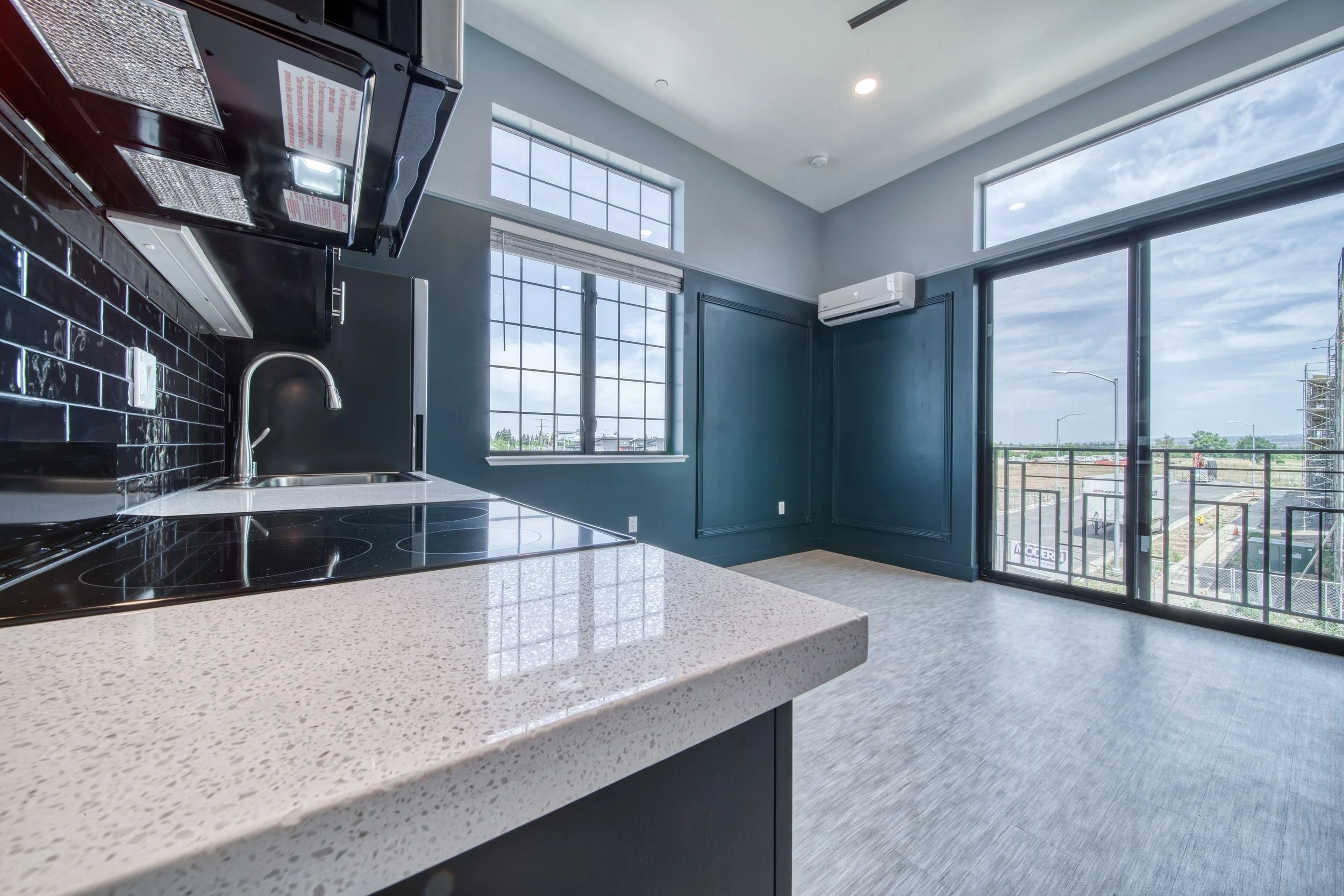 Modern kitchen with black cabinets, a black tile backsplash, a white speckled countertop, large windows, a sliding glass door leading to a balcony, and an air conditioning unit inside.
