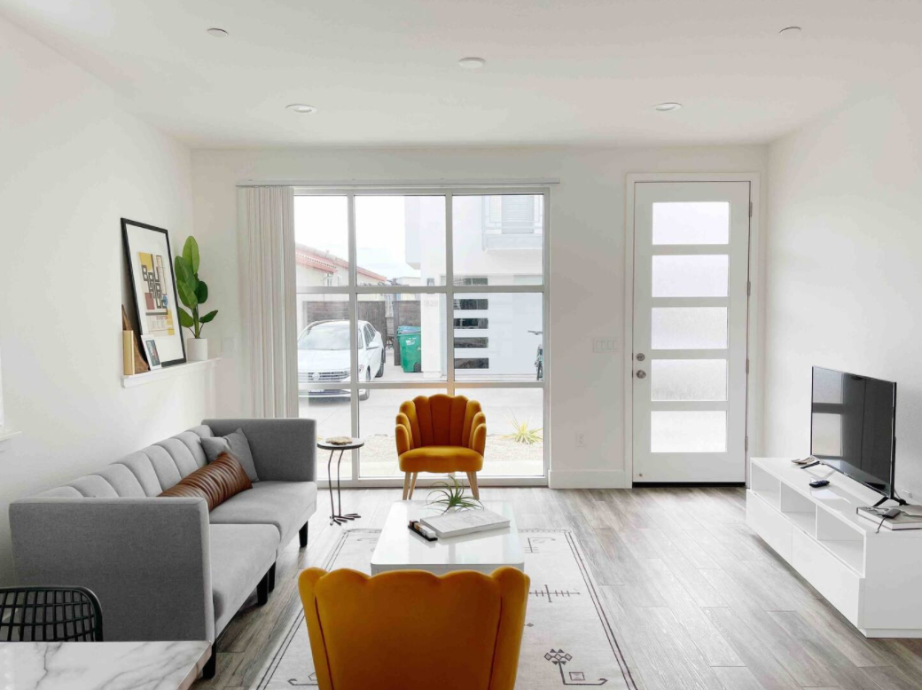 Bright living room with white walls, gray sofa, yellow accent chairs, a white coffee table, a white TV stand with a flat-screen TV, large window, and modern decor.
