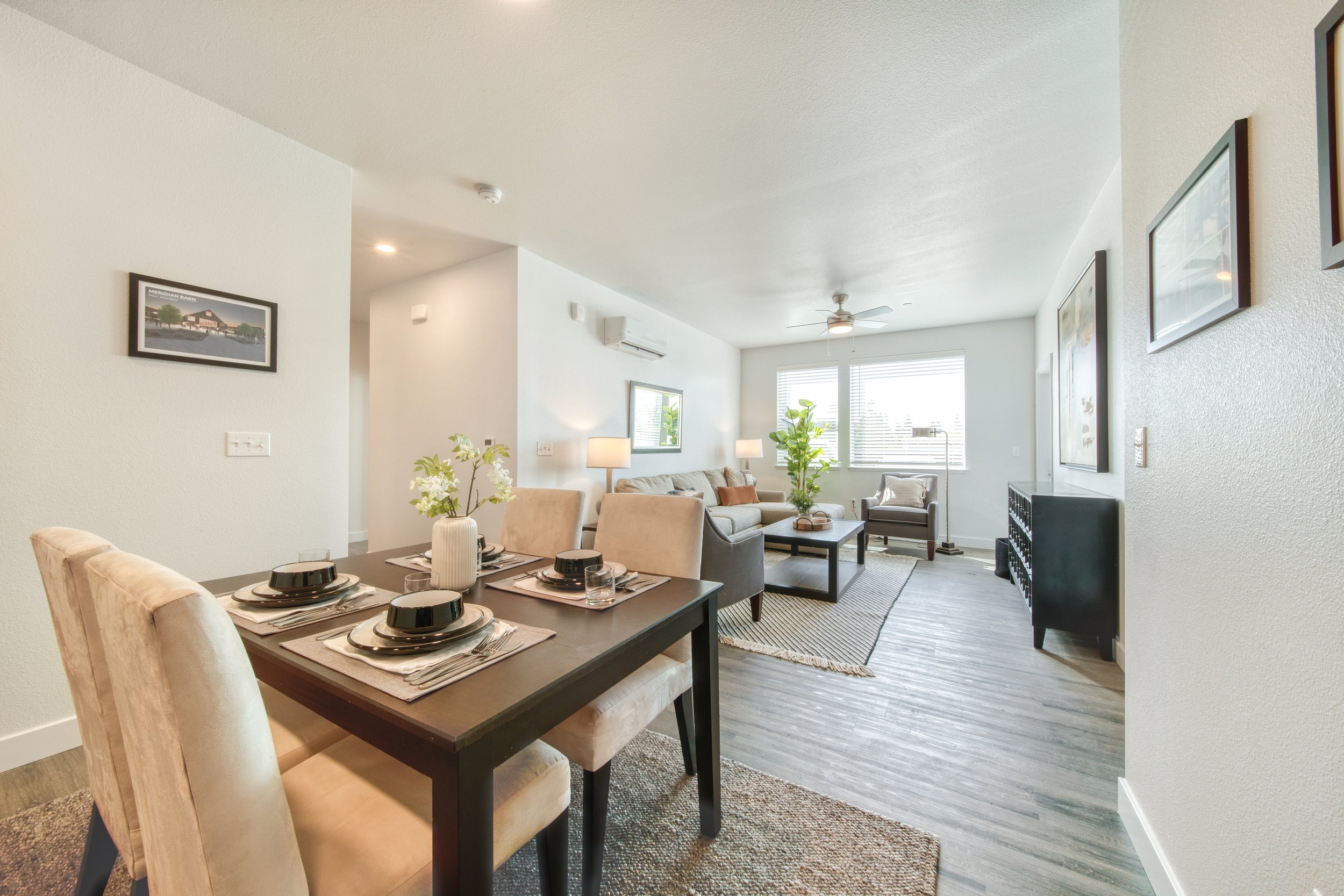 Open-concept living and dining area with modern decor, hardwood floors, beige sofa, armchair, coffee table, dining table set for four, framed artwork, and large windows with blinds