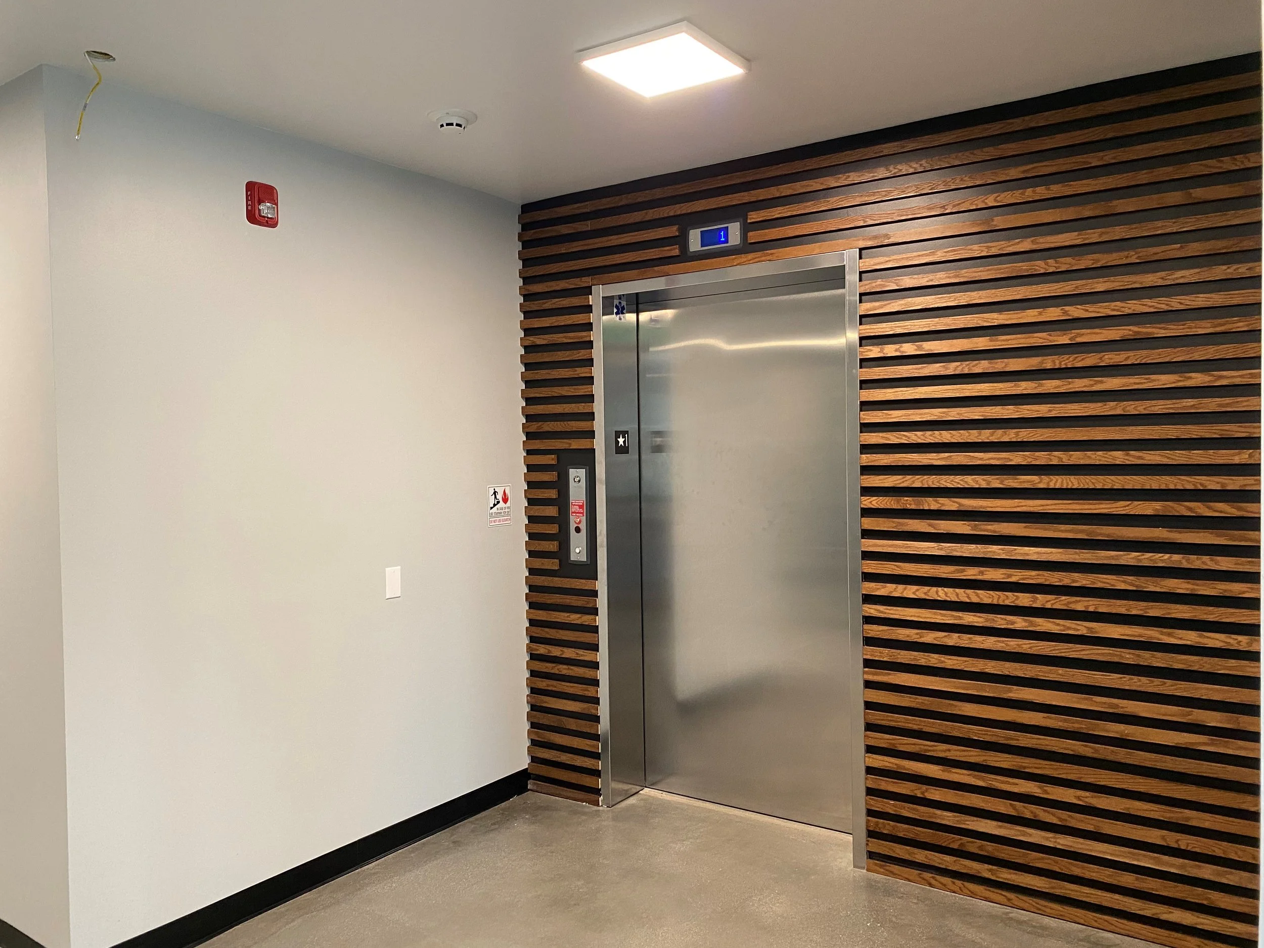 Stainless steel elevator door framed by wood paneling in a modern interior hallway.