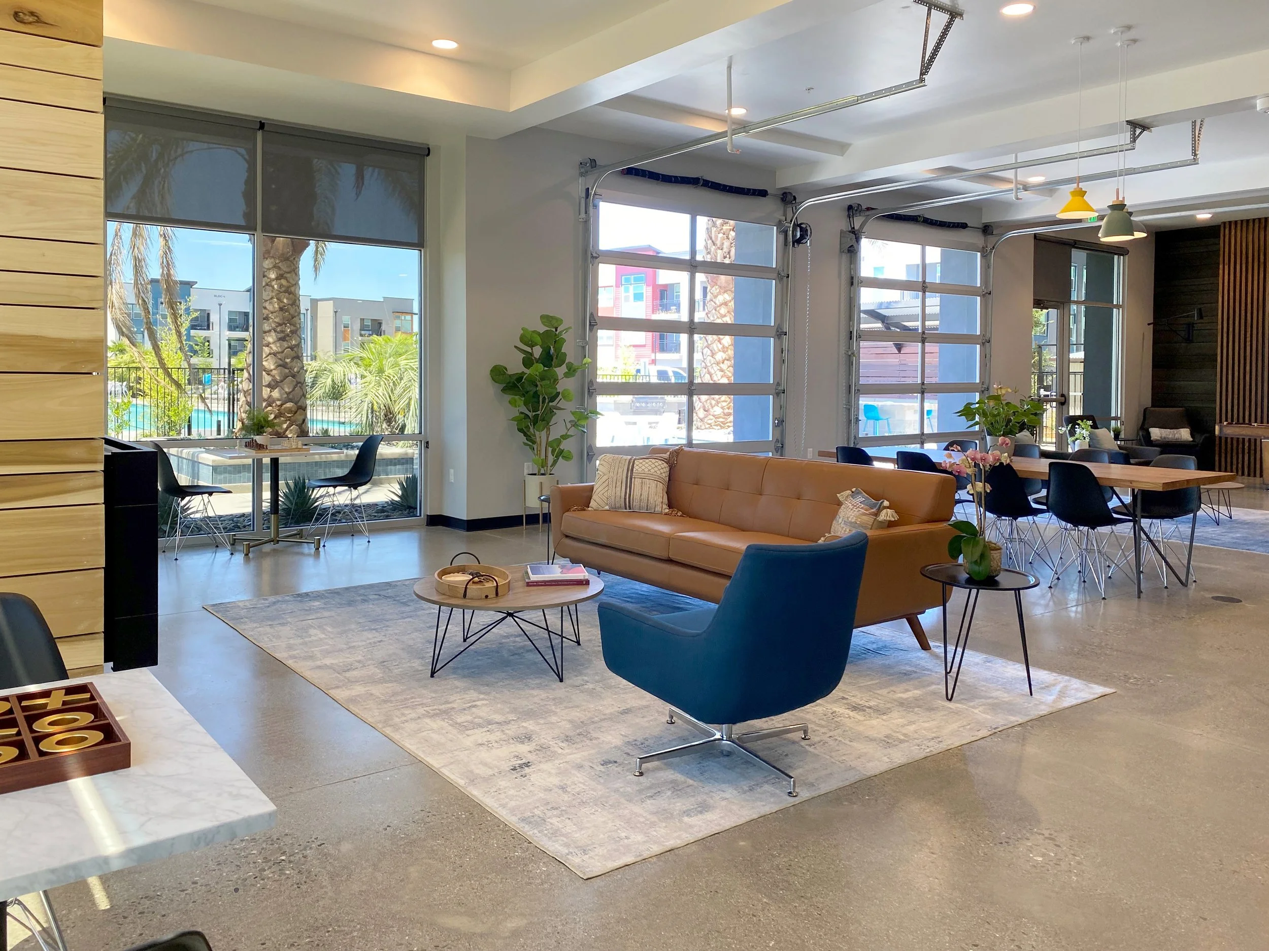 Modern, bright interior of a lounge area with large windows, a tan sofa, a blue armchair, a coffee table, and potted plants.