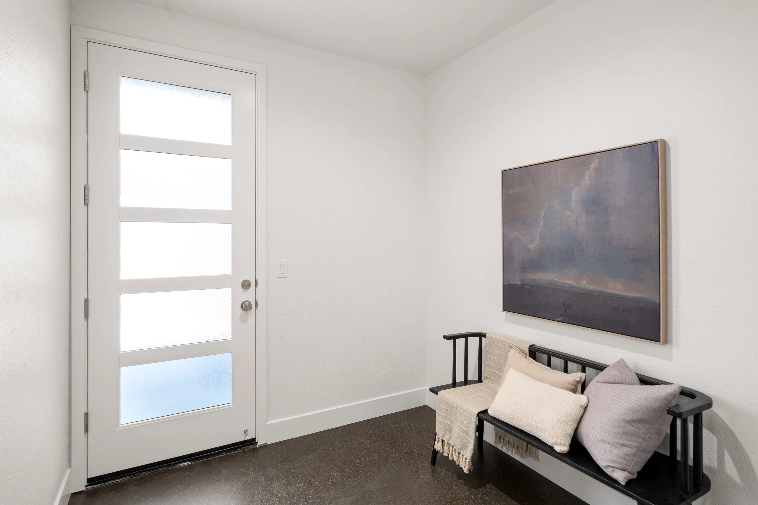 Entryway with white door, modern black bench with pillows, and wall art depicting a stormy sky.