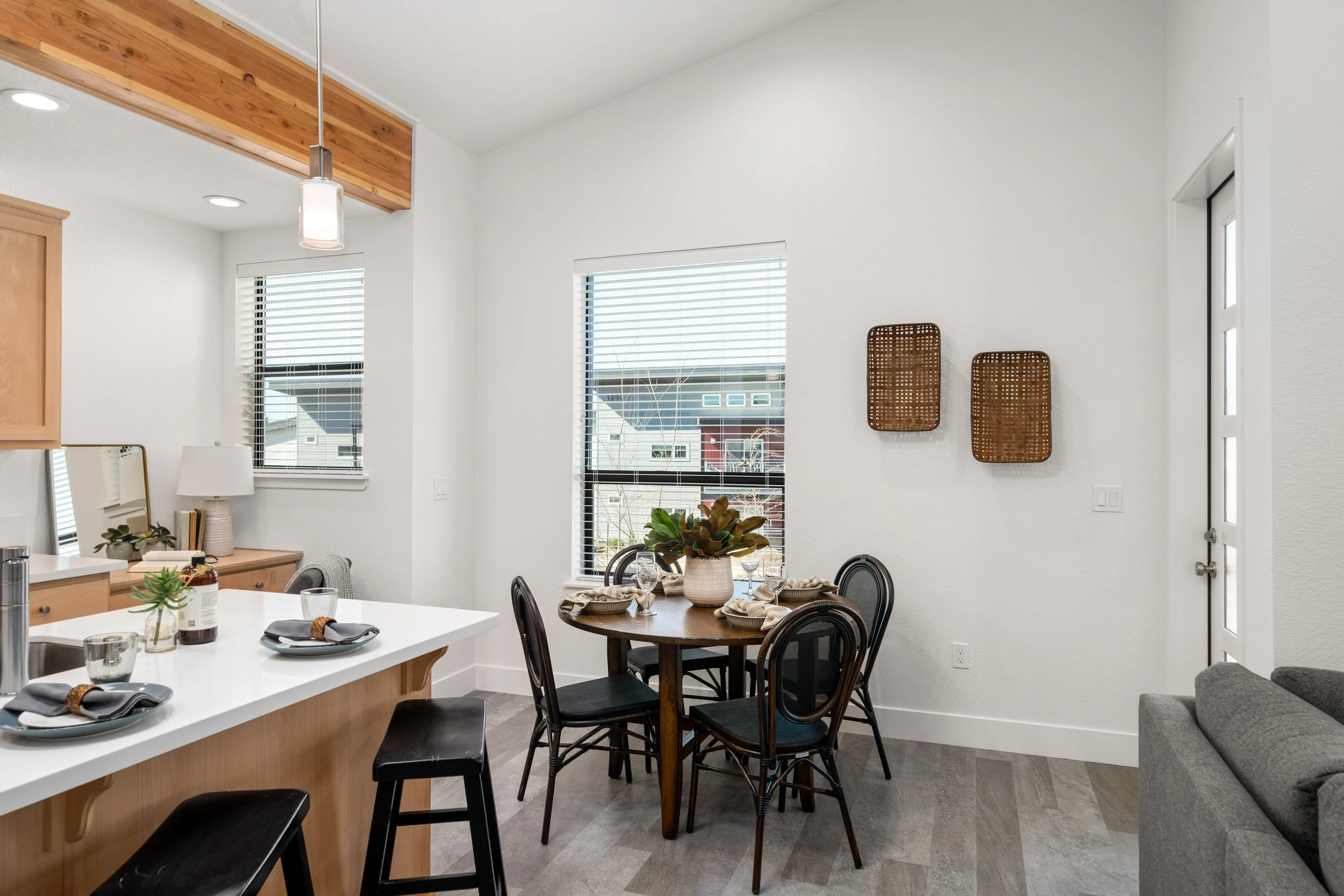 Dining area with a round wooden table set for four, decorated with a large plant centerpiece, four black chairs, adjacent to a kitchen island with stools, and windows with white blinds.
