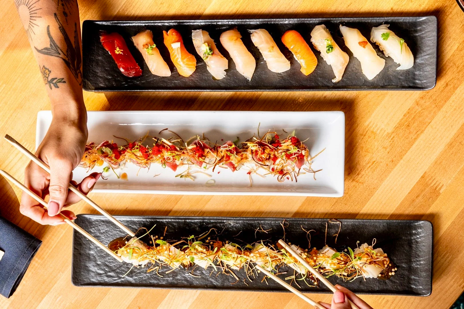 Overhead view of three rectangular plates with assorted sushi on a wooden table, with hands using chopsticks to pick up sashimi.