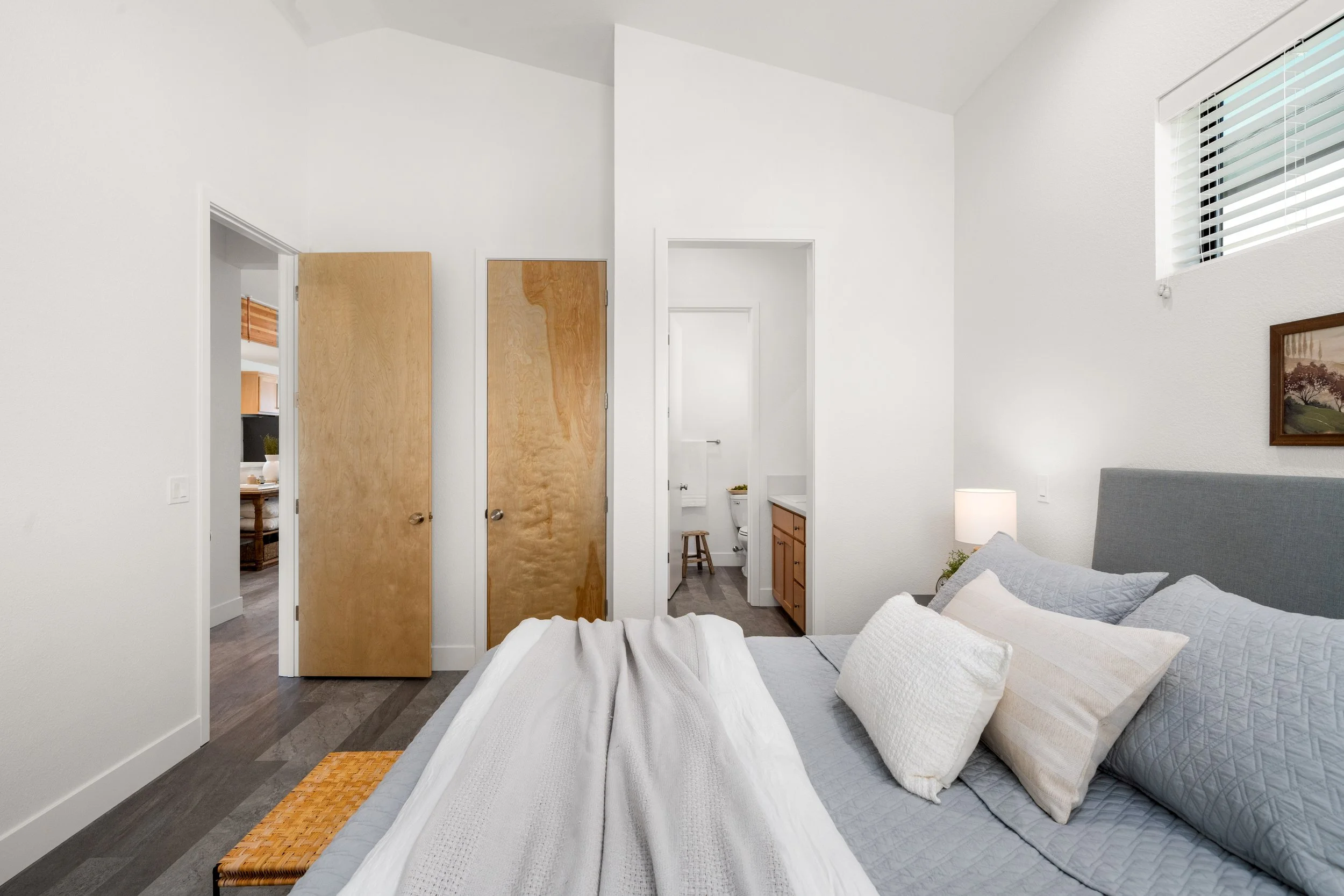A bedroom with a bed on the right, pillows, a lamp, and a window. Visible bathroom and closet doors in the background.