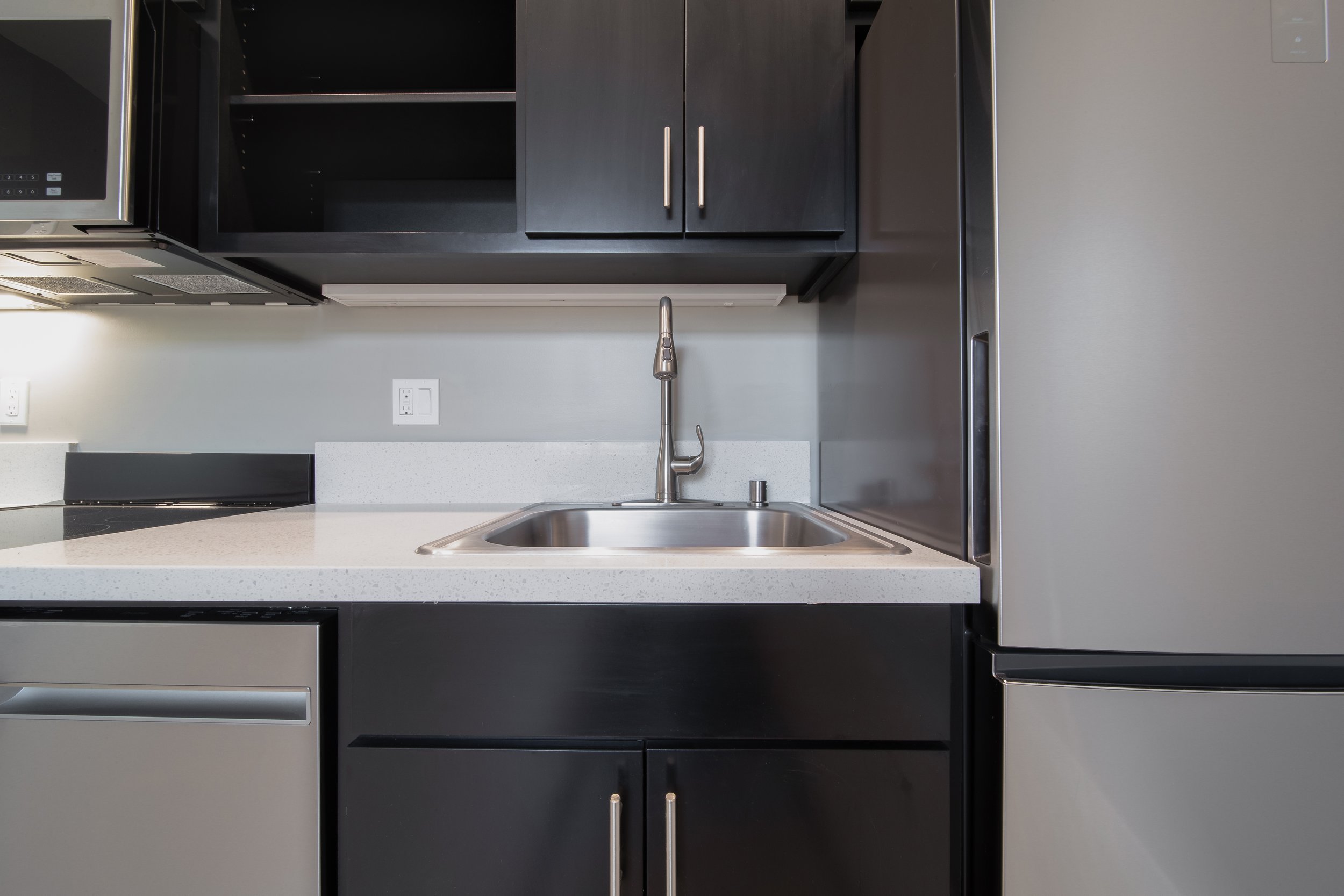Kitchen with black cabinets, a stainless steel microwave, a white countertop sink with a silver faucet, and a fridge on the right.