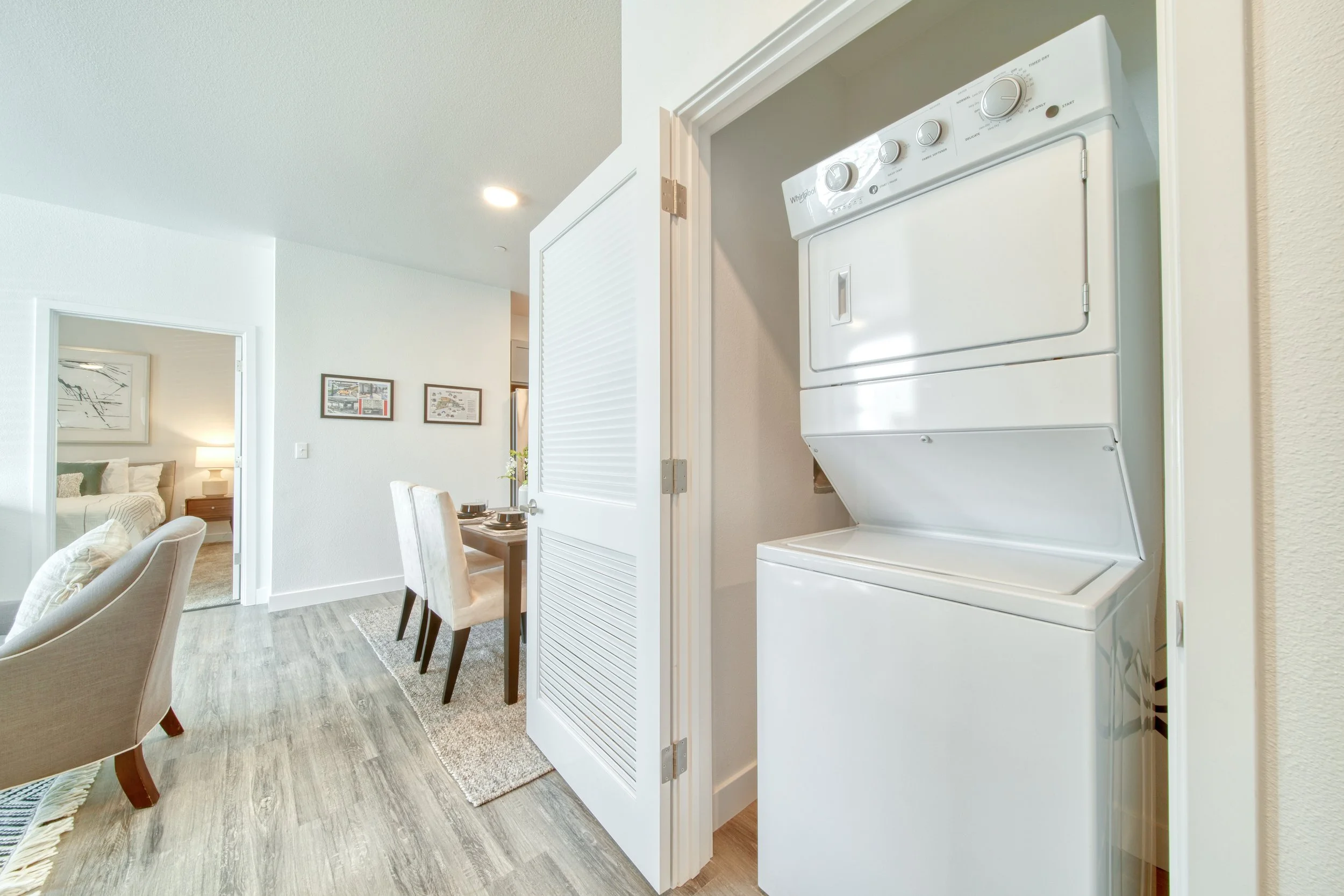 A stacked white washer and dryer inside a small laundry closet next to a dining area with a table and chairs in an open-concept living space.