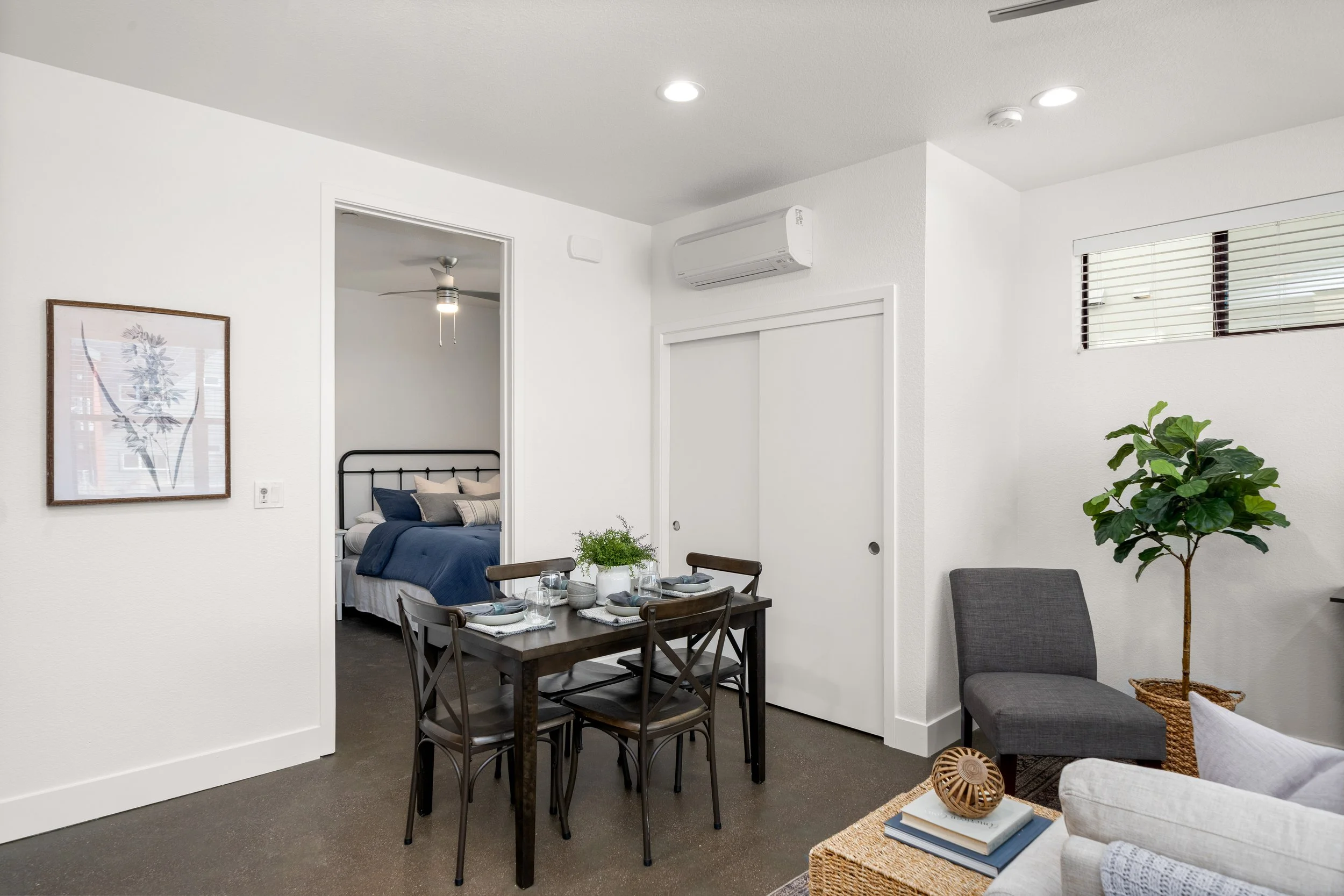 Interior of a modern living and dining area with white walls, a black dining table set for four, a sitting area with a gray chair, a TV stand, and a bedroom visible through an open door.