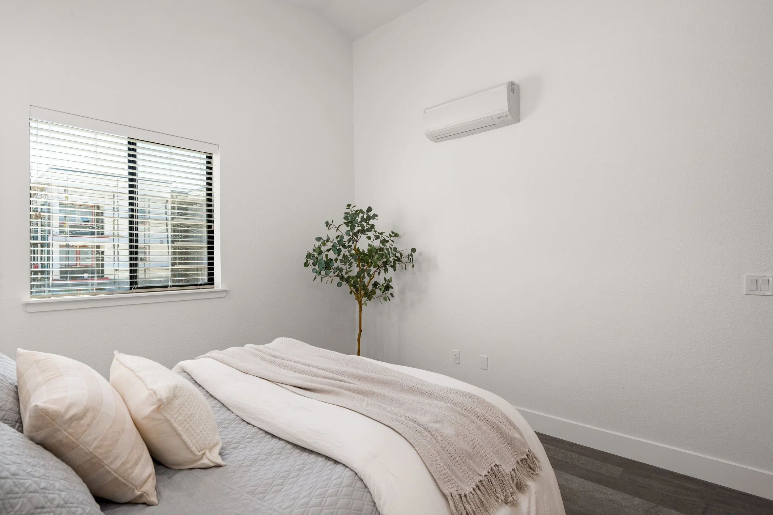 A bedroom with a bed covered in gray and white bedding, beige pillows, and a beige throw blanket. There is a window with white blinds on the left wall, and a small green plant in the corner. A white wall-mounted air conditioner is on the upper right 