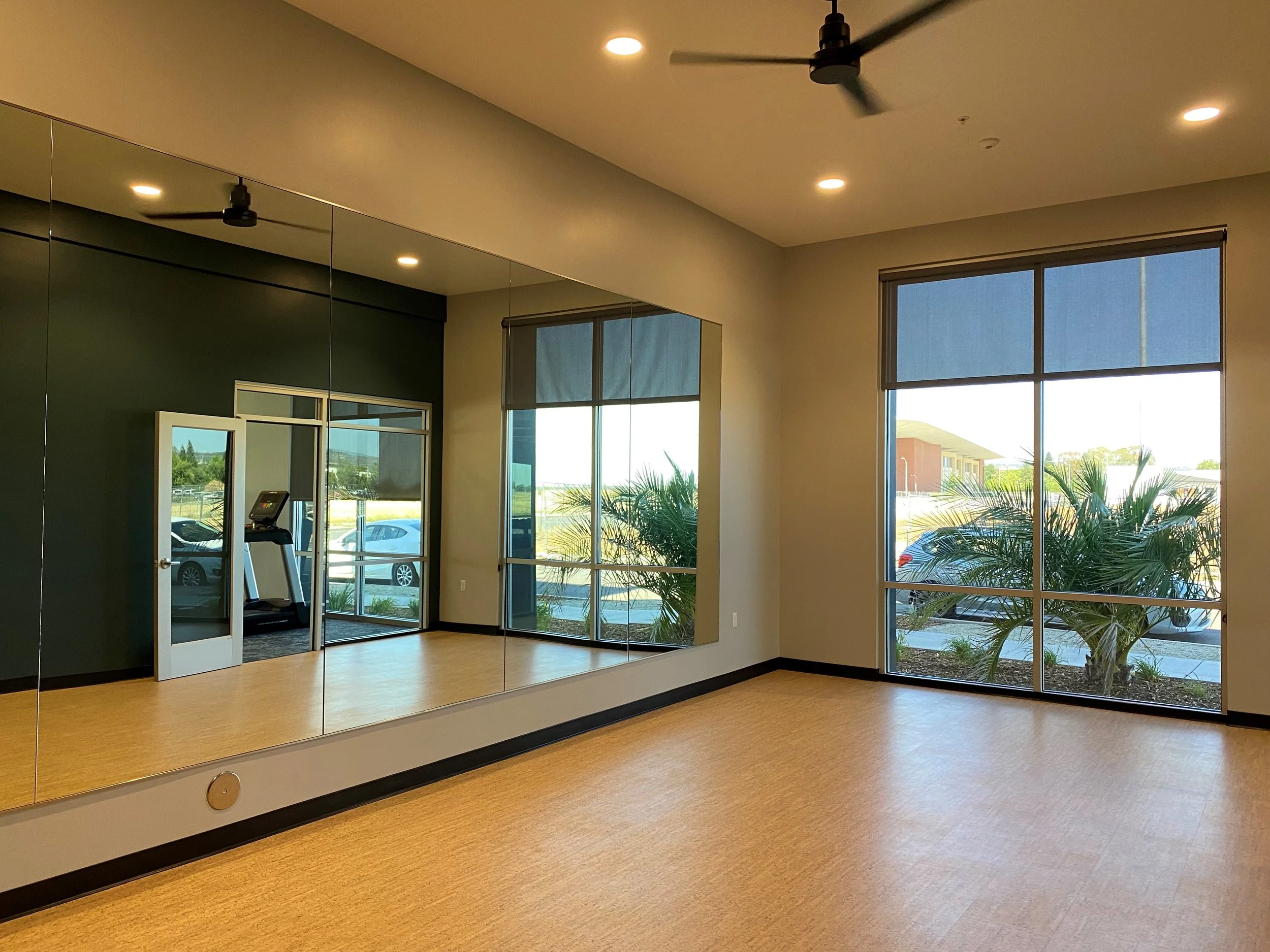 Empty room with large windows, a wall mirror, a treadmill in a small adjacent room, and ceiling fans with lights.