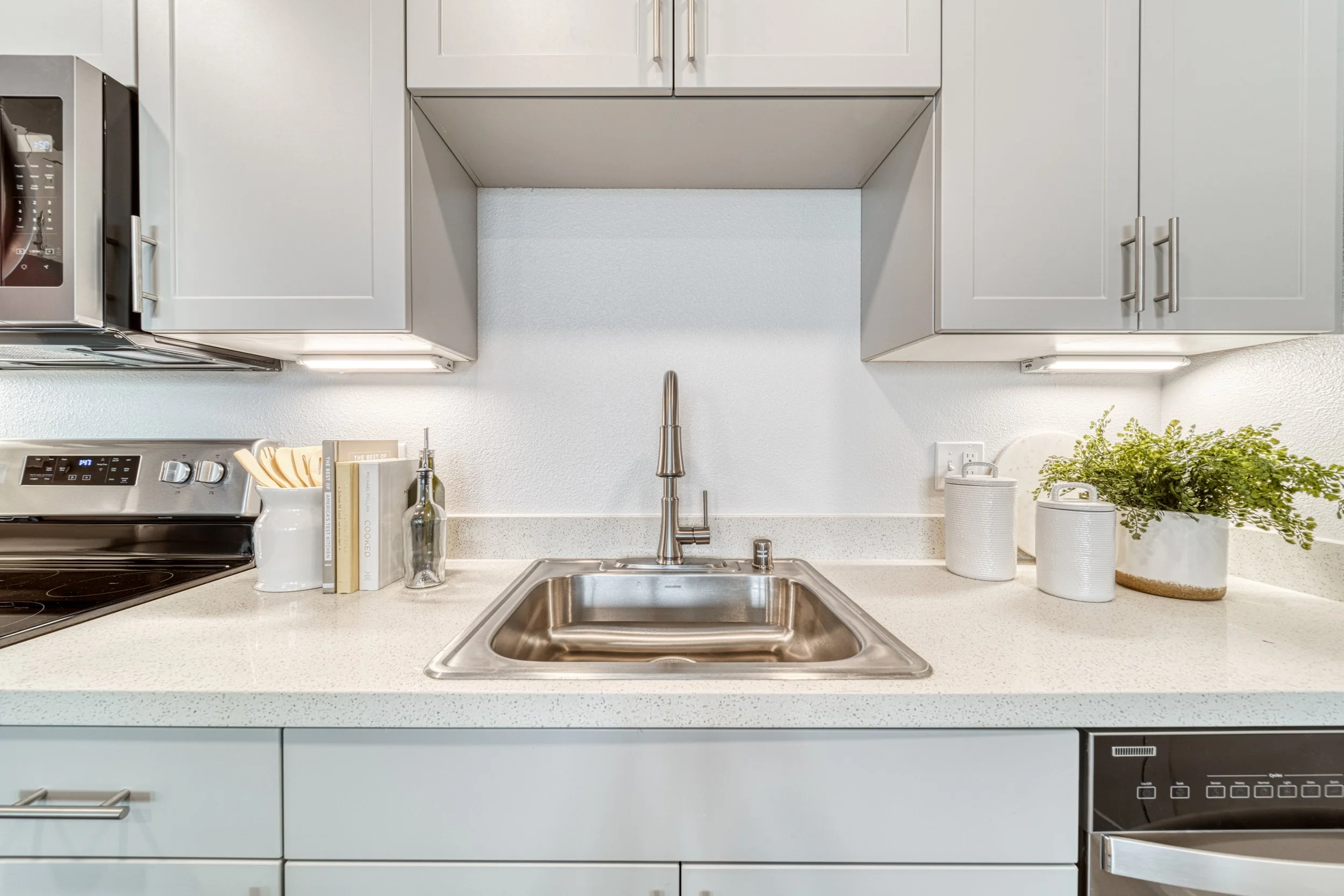 Kitchen sink area with gray cabinets, a stainless steel sink and faucet, white countertops, a microwave oven, a few books, a soap dispenser, and some potted plants.