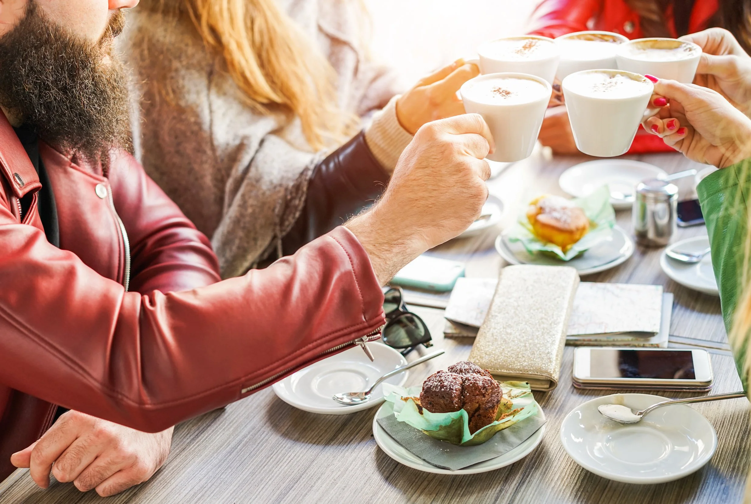 A group of people raising coffee cups in a toast during a social gathering at a restaurant or cafe, with desserts and small plates on the table.