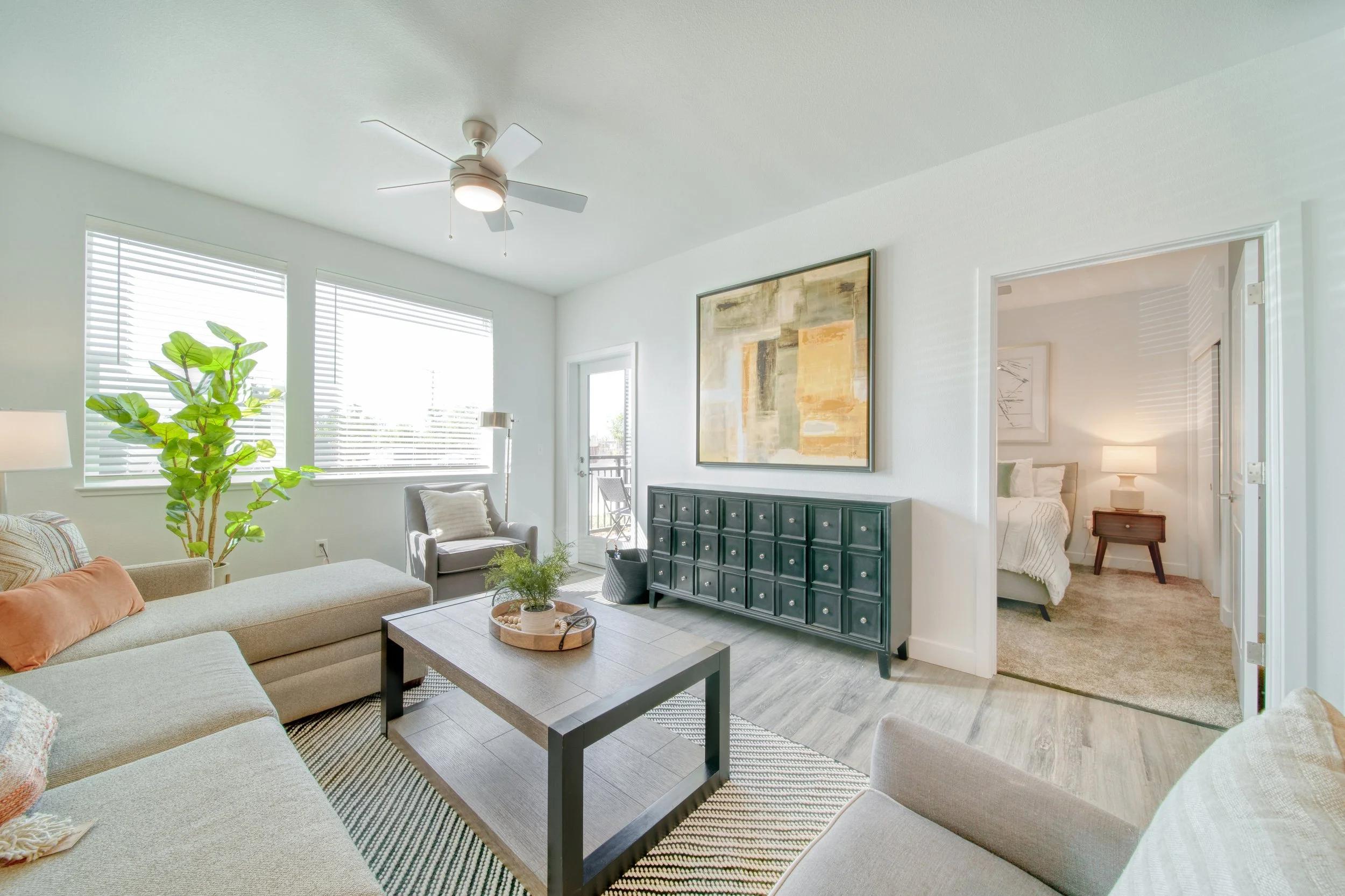 Bright living room with beige sofa, armchair, black coffee table, large window with blinds, and a sliding door leading to a balcony. A green plant is near the window, and the walls are decorated with abstract art. A bedroom with a bed and nightstand 