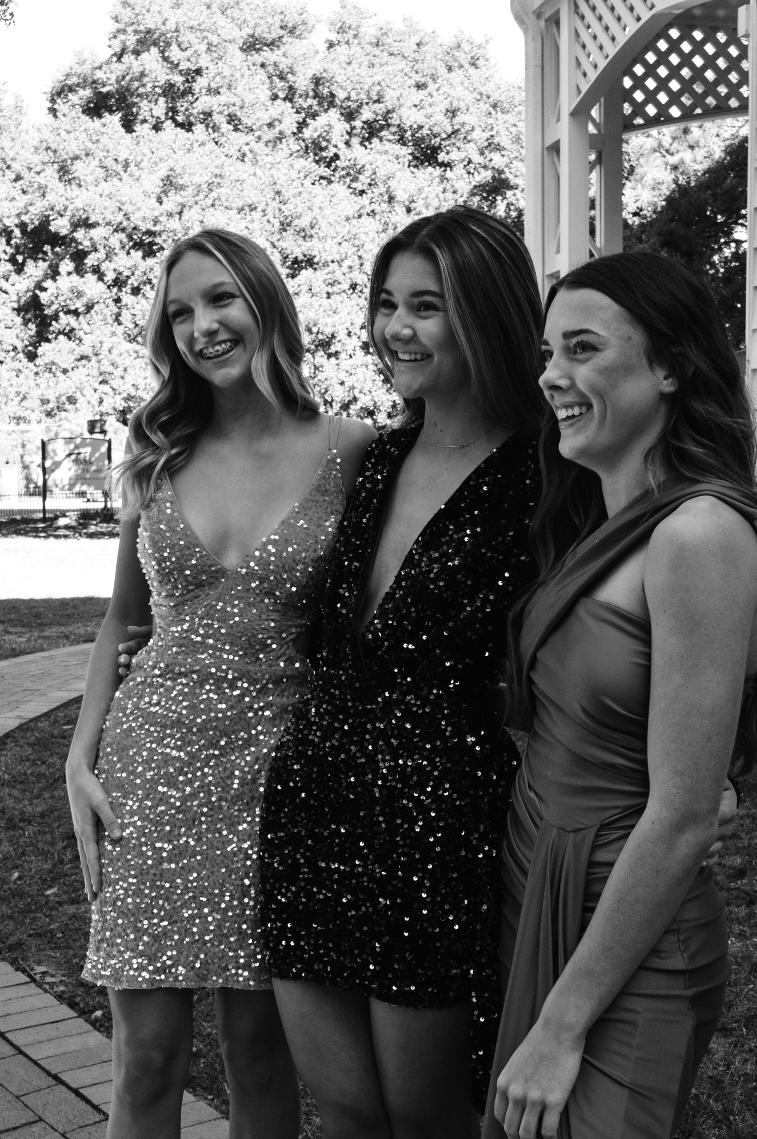 Three young women dressed in formal attire, smiling and posing together outdoors. Two are wearing sparkly dresses, and the third is in a satin gown. They are standing on a brick pathway with trees and a white gazebo in the background.