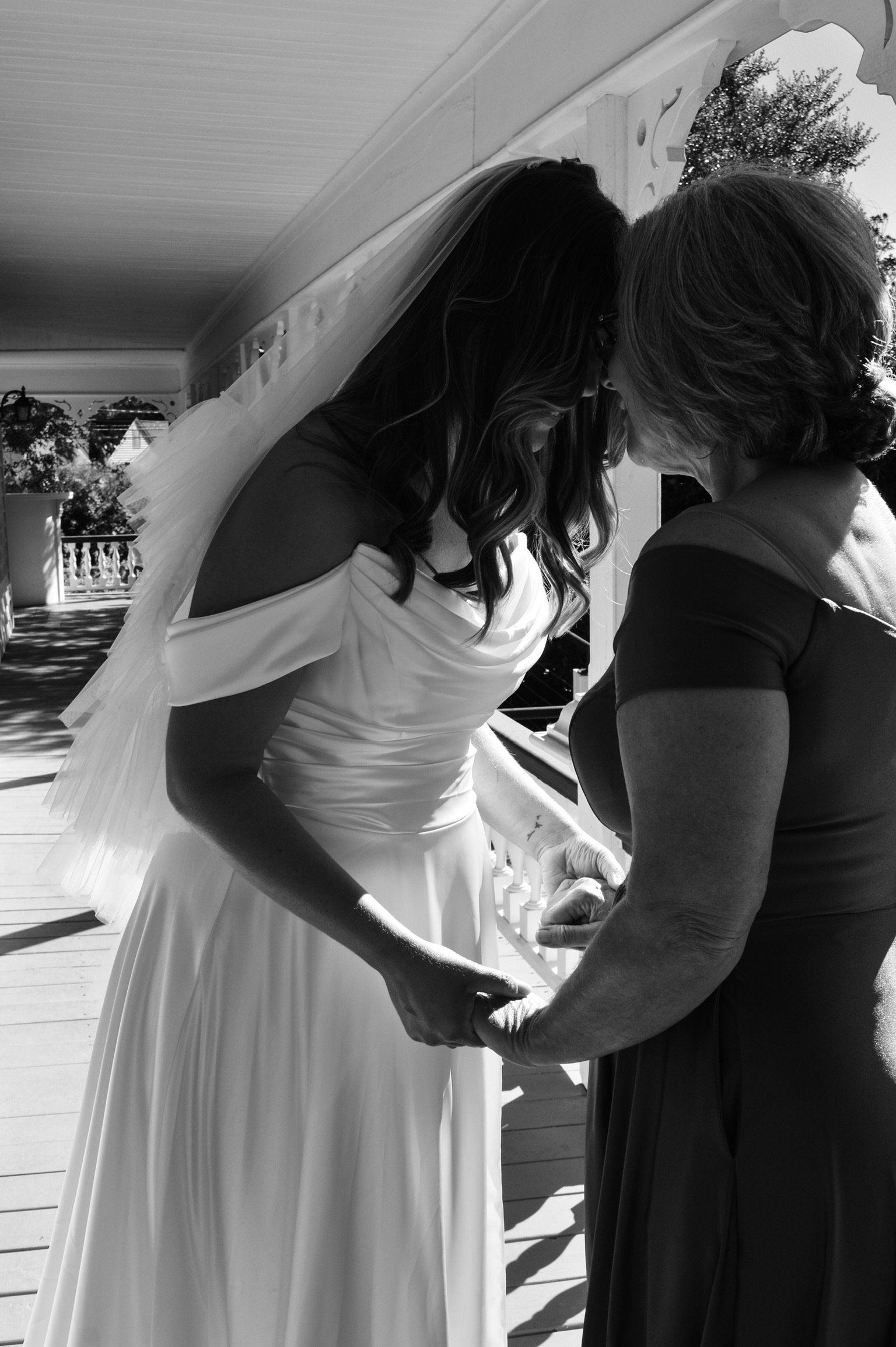 A bride in a wedding dress holding hands with her mother, on a porch during a sunny day.