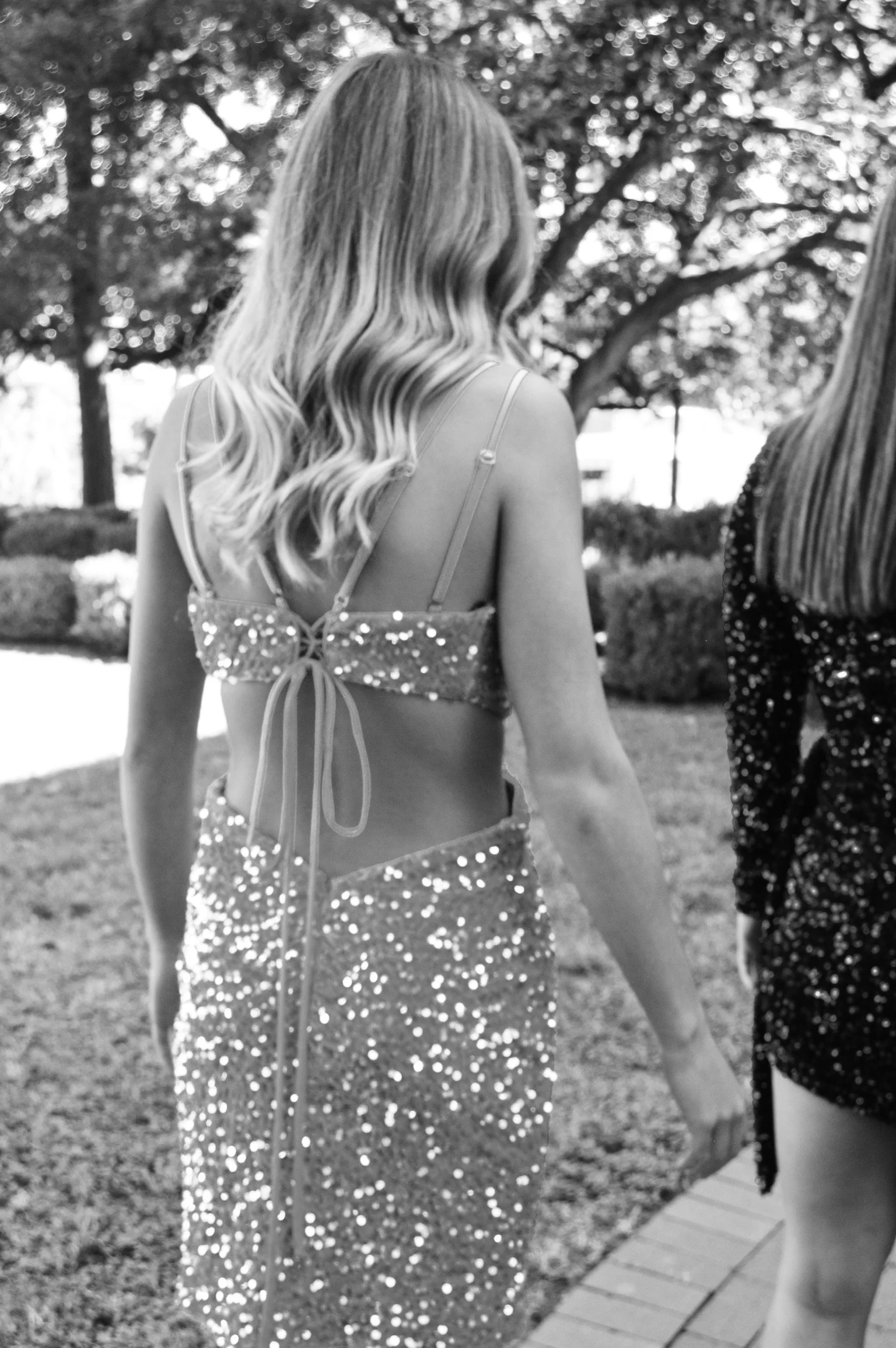 A woman in a sparkly, sequined dress with a open back, walking outdoors with trees and bushes in the background.