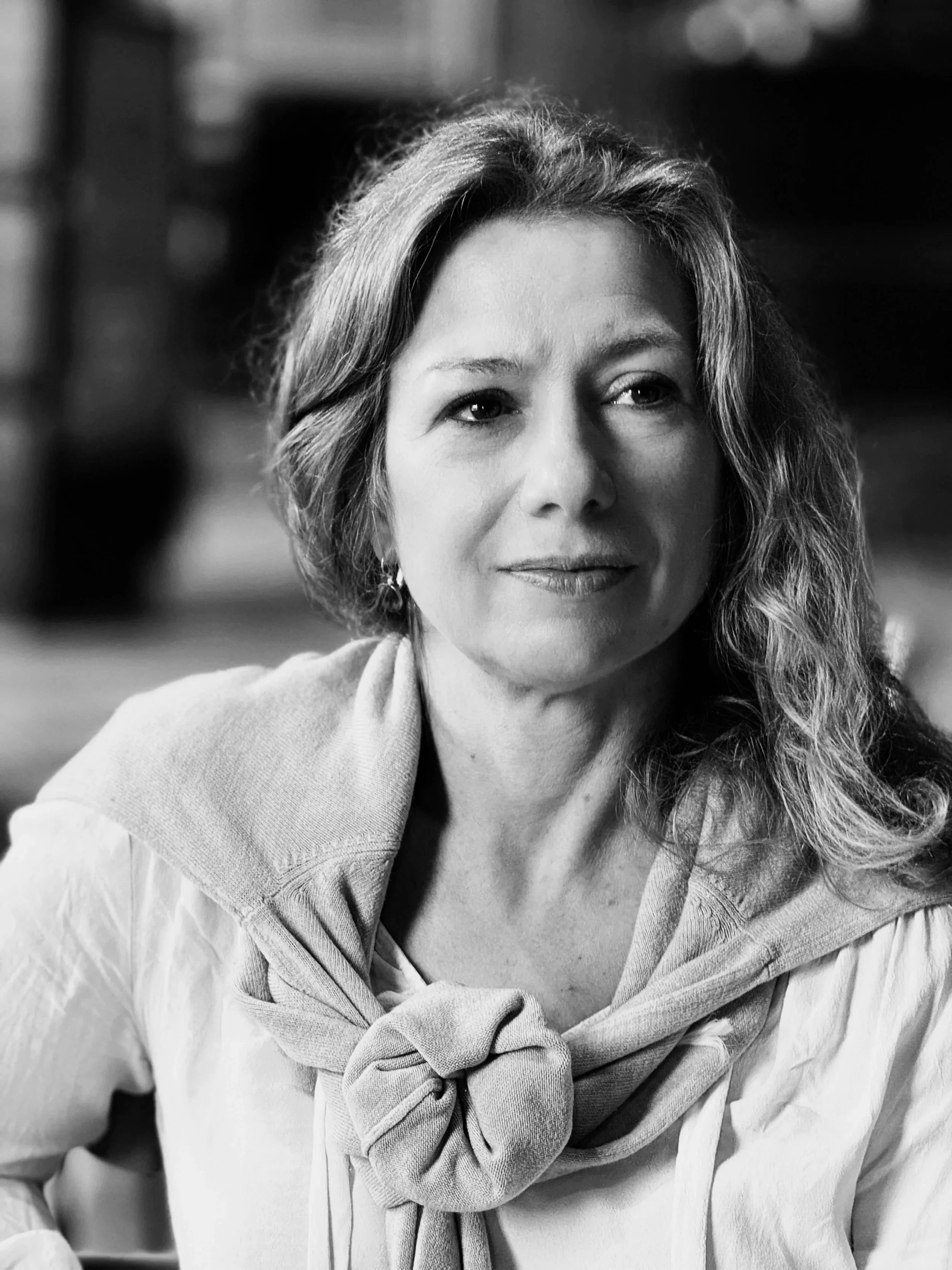 Black and white photograph of a woman with curly hair, wearing a light-colored shirt and scarf, smiling softly, with an outdoor background.