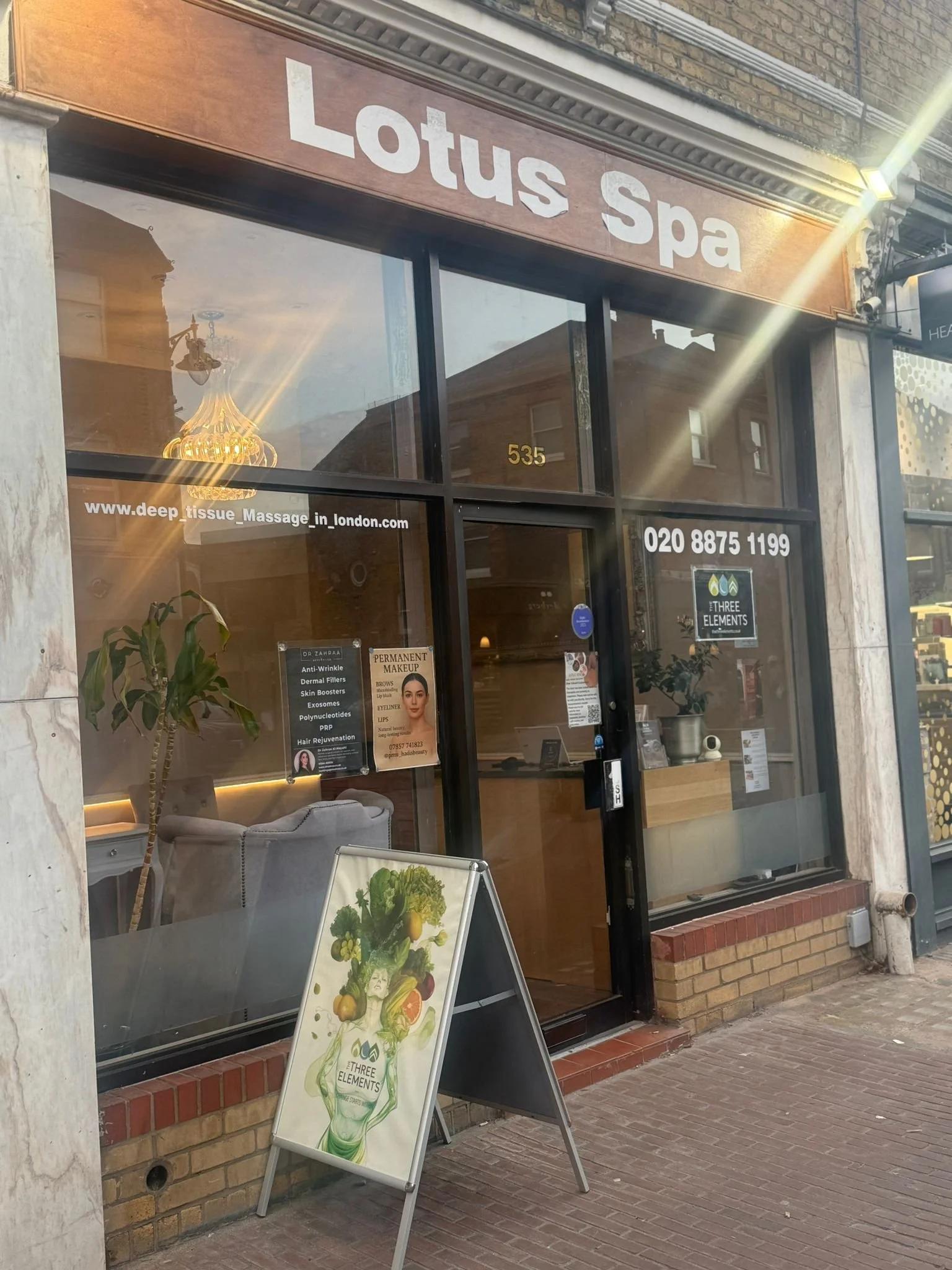 storefront of Lotus Spa with large glass windows and an orange sign. There is an A-frame sign outside displaying a graphic of a woman with vegetables and the spa's name, and a potted plant near the entrance. The business offers massage and skincare services.