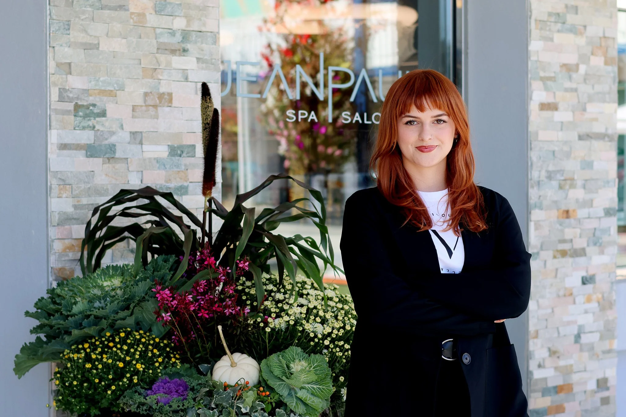 A woman with red hair stands in front of a spa and salon. She is wearing a black jacket over a white shirt and has her arms crossed. There are colorful flowers and a white pumpkin beside her.