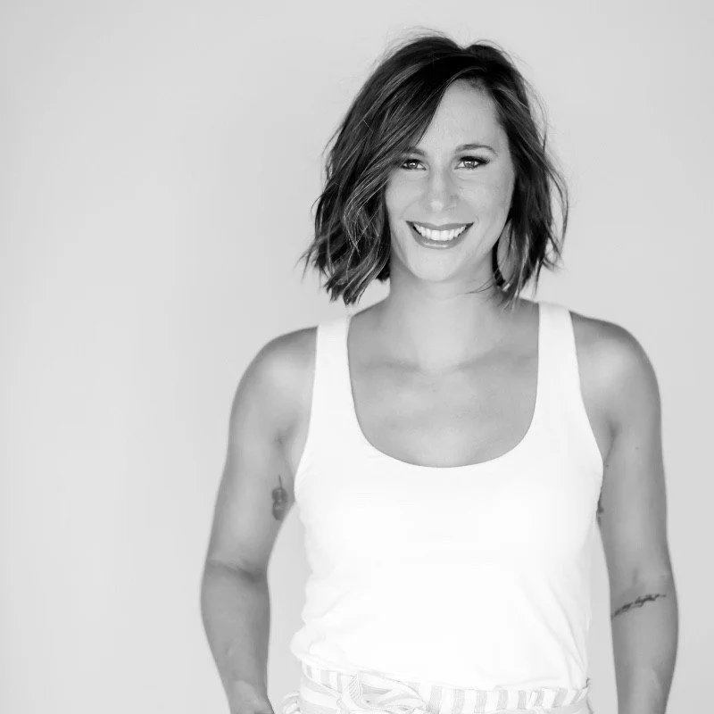 Black and white photo of a woman with a bob haircut, smiling, wearing a white tank top and striped pants, standing against a plain background.