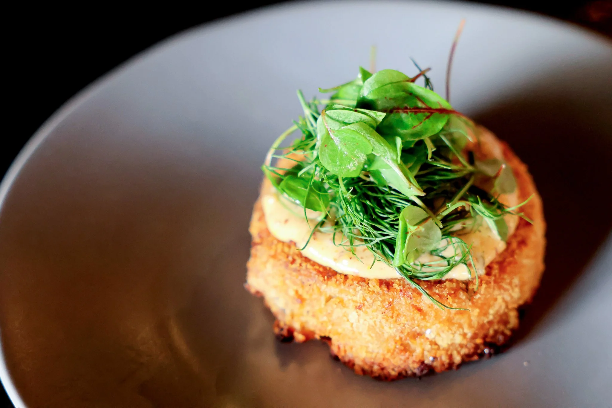 A plated dish featuring a crispy breaded piece topped with fresh mixed greens and herbs, served on a dark plate.