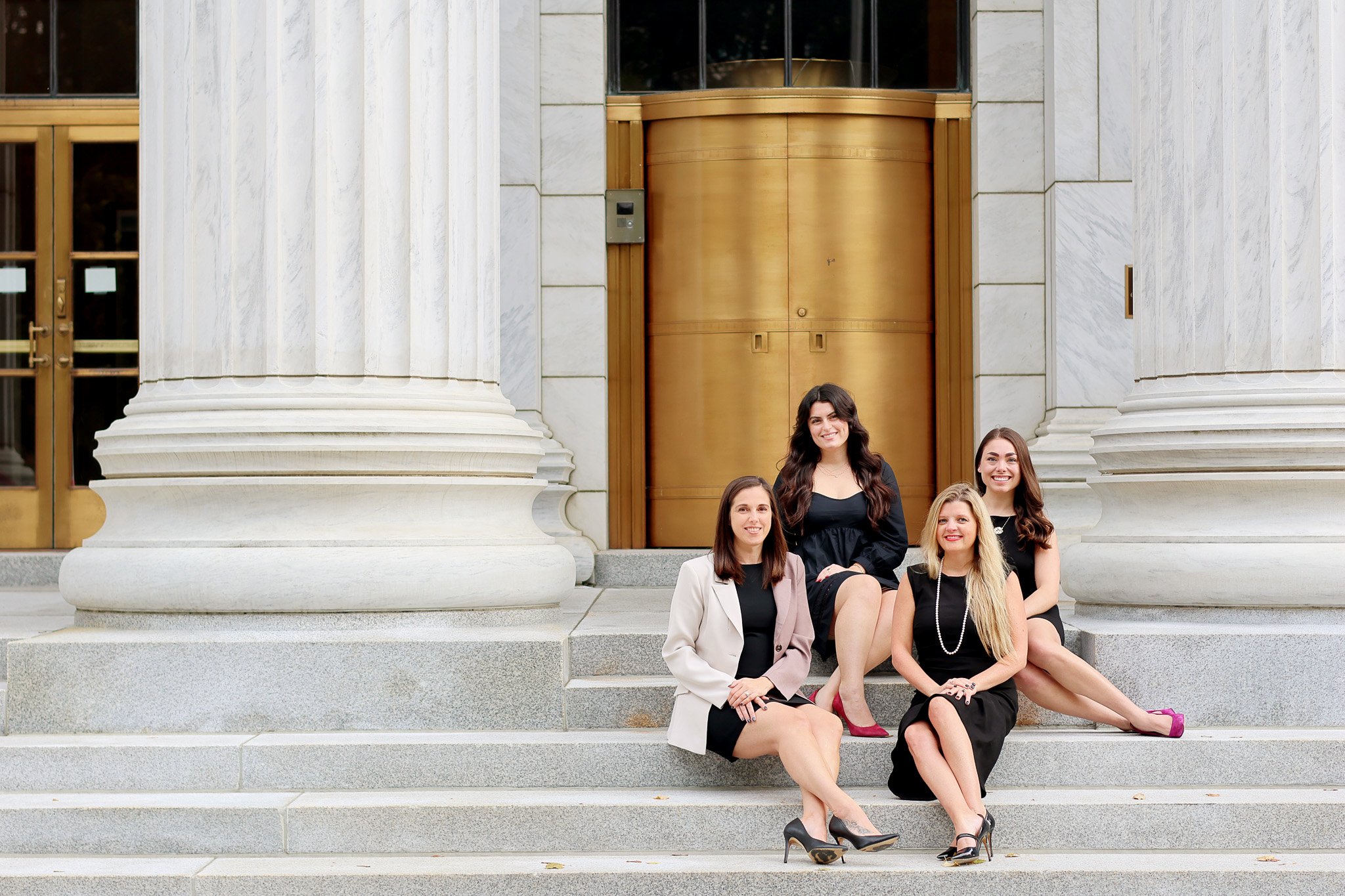 Four women sitting on front stairs of a building with large white columns and a wooden door behind them, smiling.