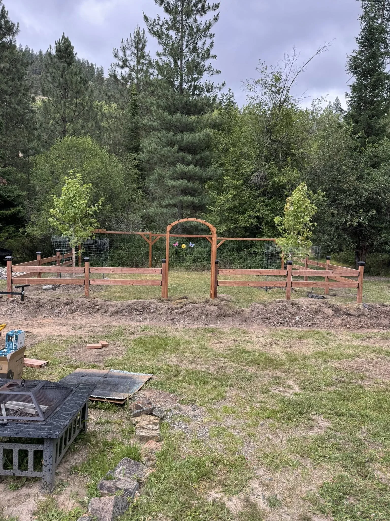 Backyard with a fenced garden area, wooden gate, surrounded by trees, with construction materials and tools nearby.