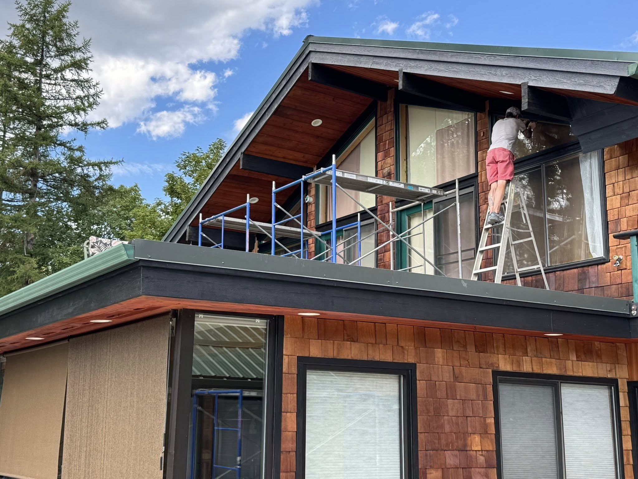 A man in a gray t-shirt and red shorts is standing on a ladder working on a window of a two-story house with brown wooden siding and large glass windows. The house has a green roof and a small balcony. The sky is cloudy with patches of blue, and there are tall trees in the background.