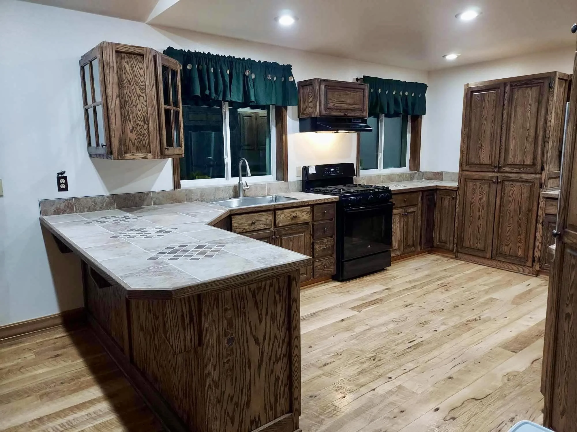 A kitchen with wooden cabinets, a black stove, white countertops with tile patterns, and two large windows with green curtains. The kitchen has wooden flooring and recessed ceiling lights.