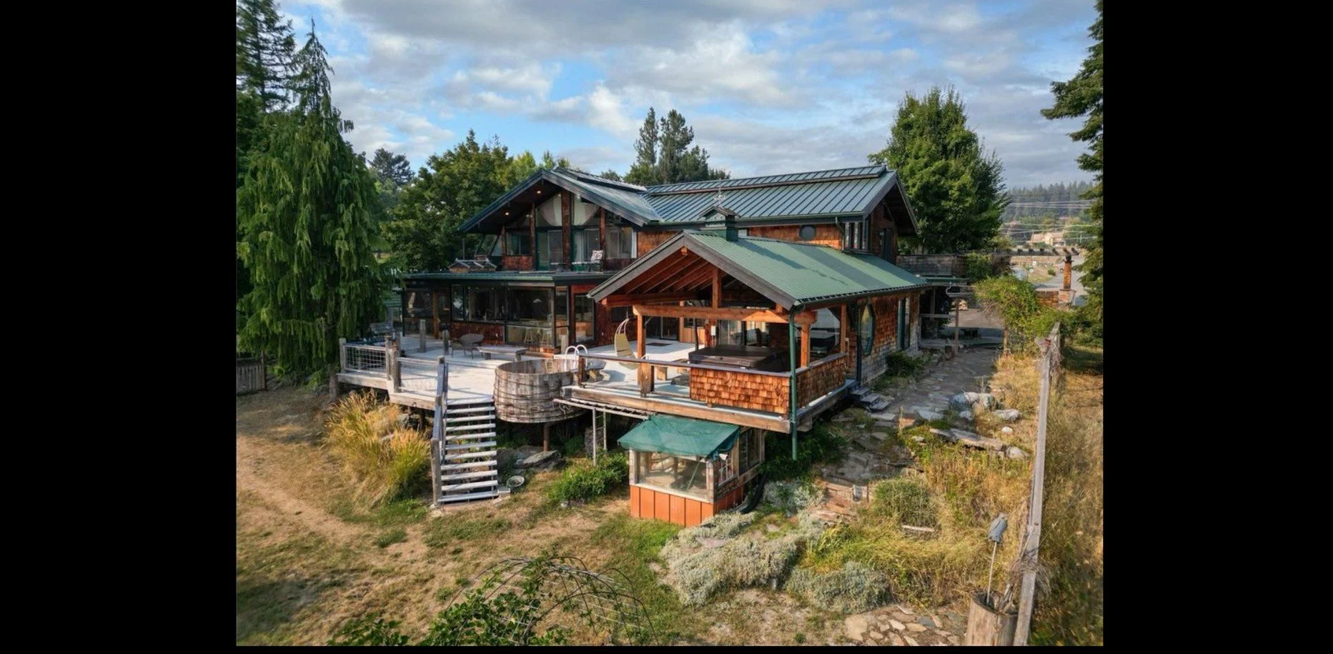 A multi-level house with large glass windows, a wooden deck, and a green metal roof, situated on a slope with trees and a partly cloudy sky in the background.