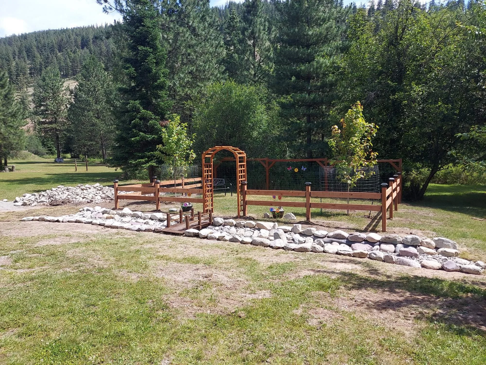 A landscaped yard with a wooden garden fence, decorative archway, and rocks, surrounded by trees and mountainous terrain in the background.