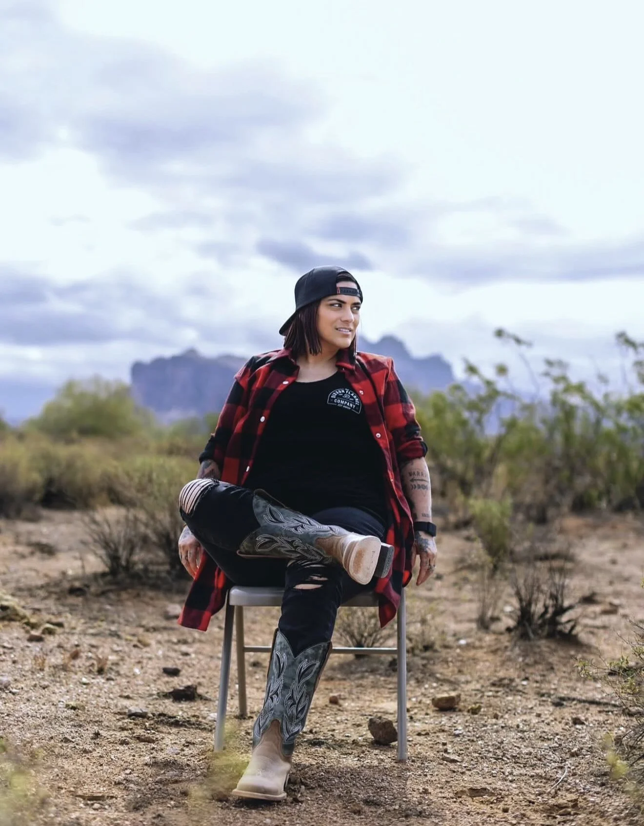 A woman in western wear sitting on a chair outdoors in a desert landscape with mountains and clouds in the background.
