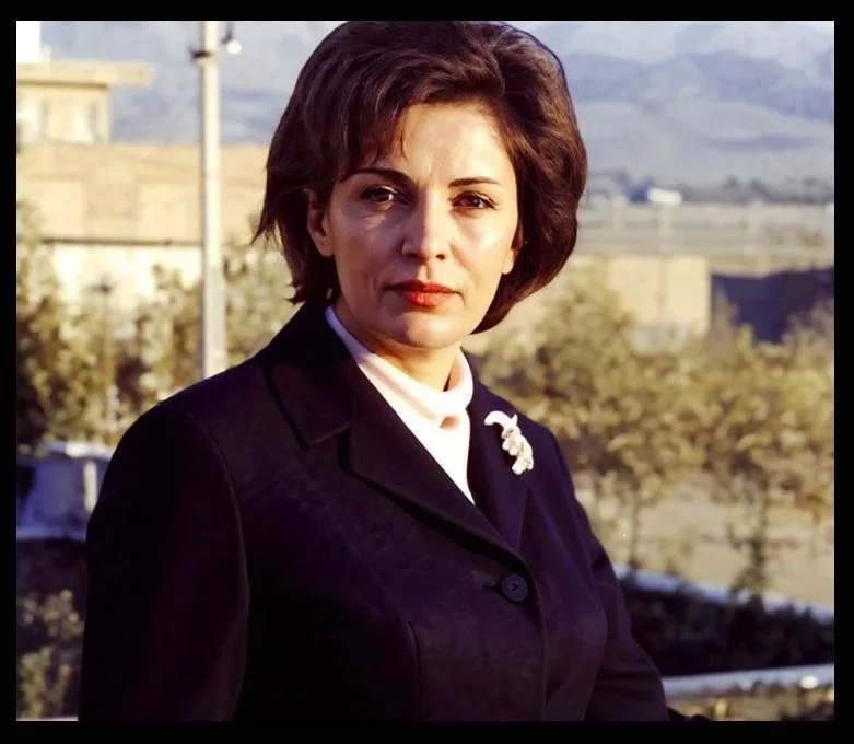 A woman with short brown hair wearing a dark blazer with a brooch, standing outdoors with a blurred background of trees and buildings.