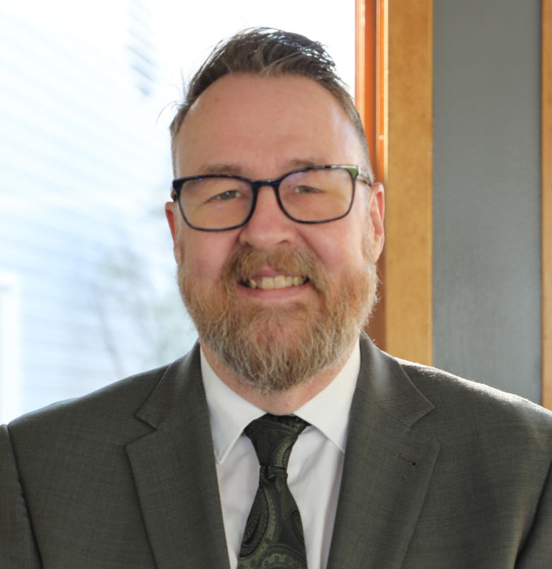 A middle-aged man with glasses, a beard, and a confident smile wearing a gray suit, white shirt, and paisley tie, sitting near a window with wood trim, bright natural light in the background.