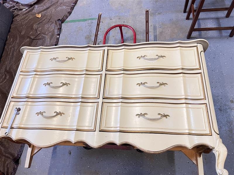 A vintage cream-colored dresser with six drawers, ornate handles, and scalloped edges, placed on a wooden support frame in a workshop or furniture refinishing area.
