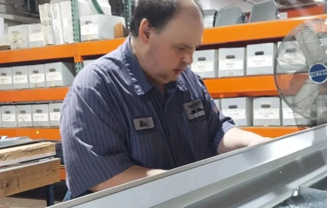 Man working in a warehouse or storage area, wearing a blue uniform, surrounded by organized boxes and shelves, with an industrial fan nearby.