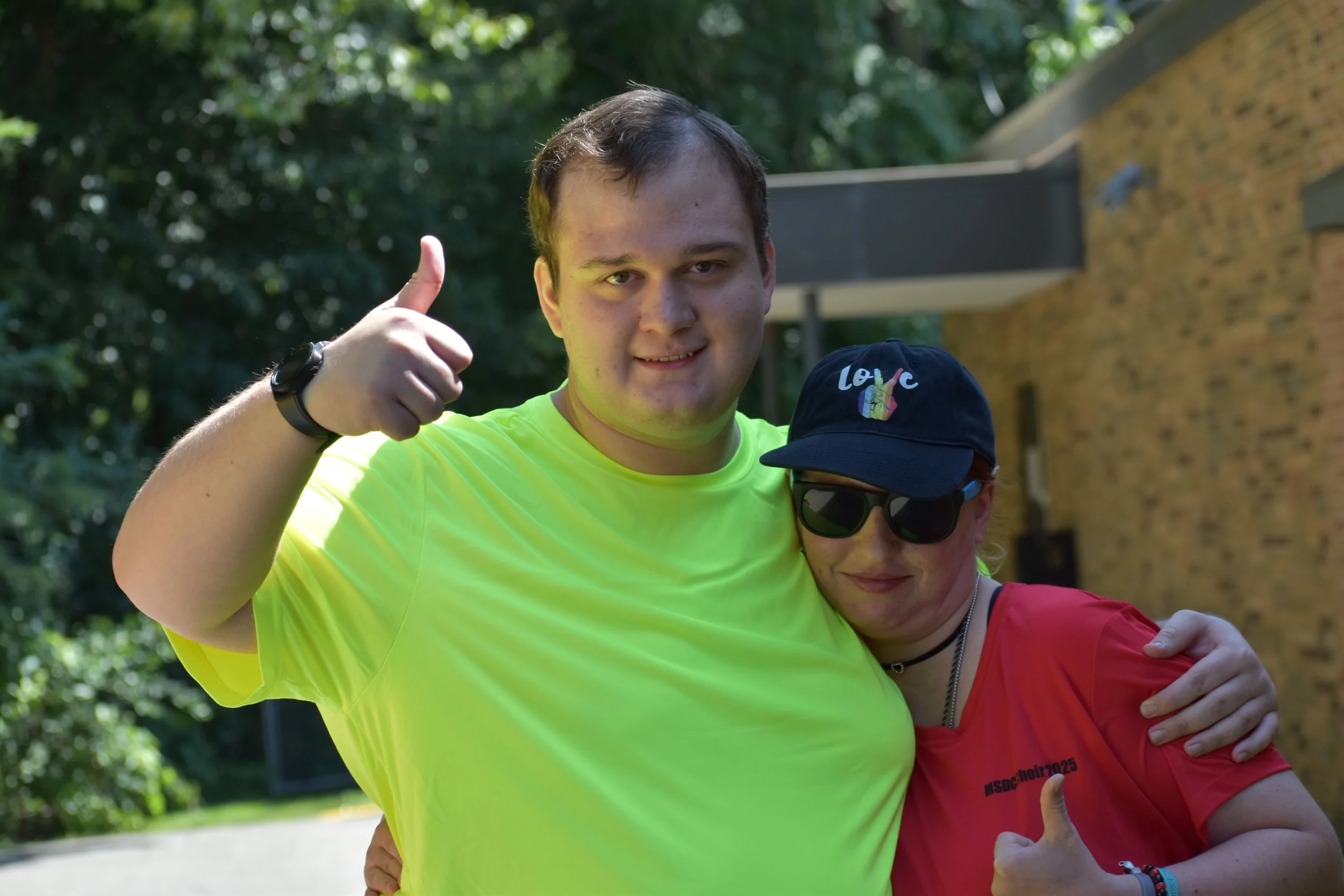 Two people hugging outdoors, one giving a thumbs up and smiling, the other wearing sunglasses and a cap with a peace sign, both appearing happy.