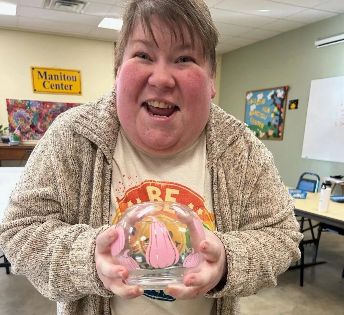 A cheerful person holding a glass paperweight with pink tulip designs inside, standing in a room with a "Manitou Center" sign and colorful artwork on the walls.