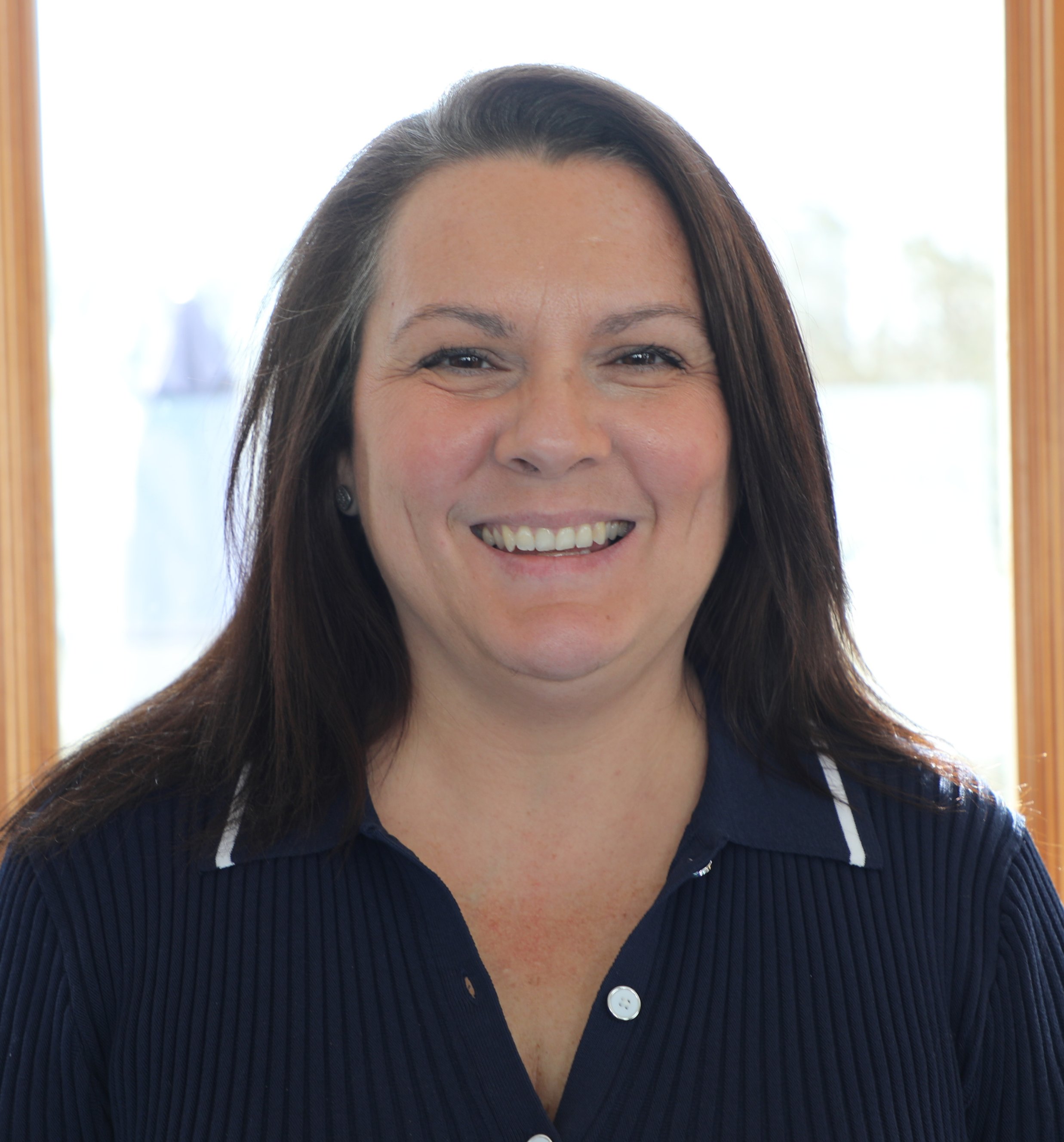 A woman smiling with shoulder-length dark hair, wearing a navy blue shirt, standing in front of a window with natural light.