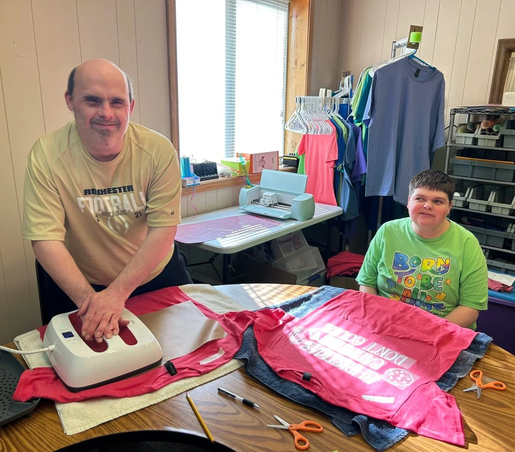 A man and a boy with disabilities at a craft table with fabric, scissors, and a heat press machine, working on a T-shirt project in a room with clothing and fabric supplies.