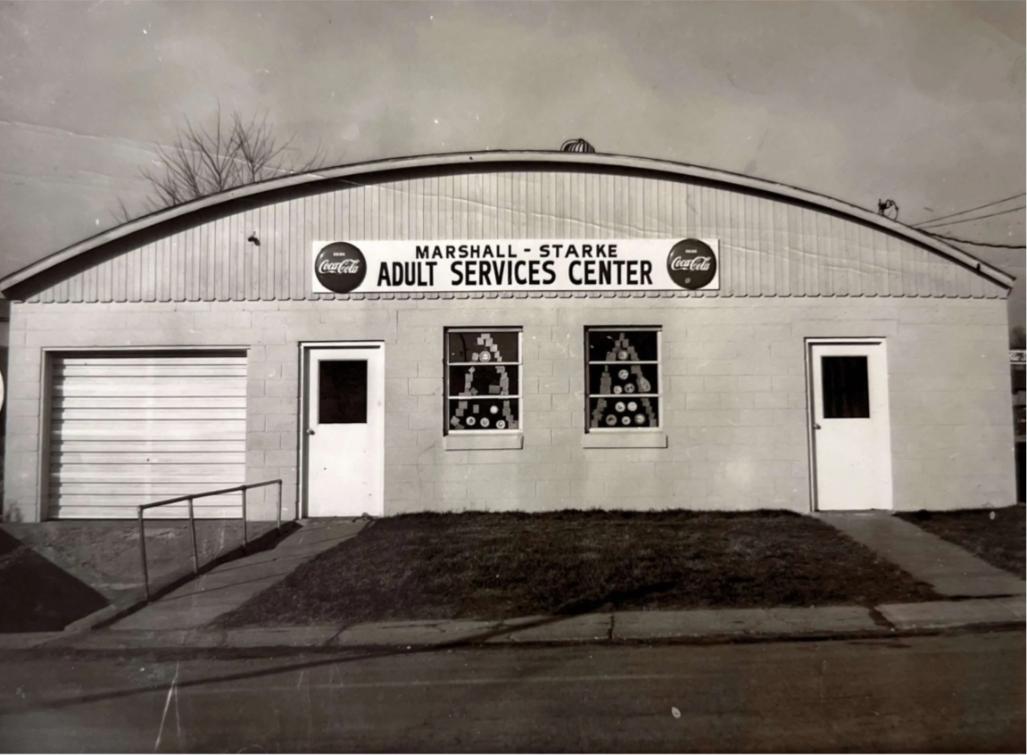 Black and white photo of a single-story building labeled 'Marshall-Starke Adult Services Center' with Coca-Cola signs above the sign. The building has two doors, three windows, a small concrete ramp, and a grassy area in front.
