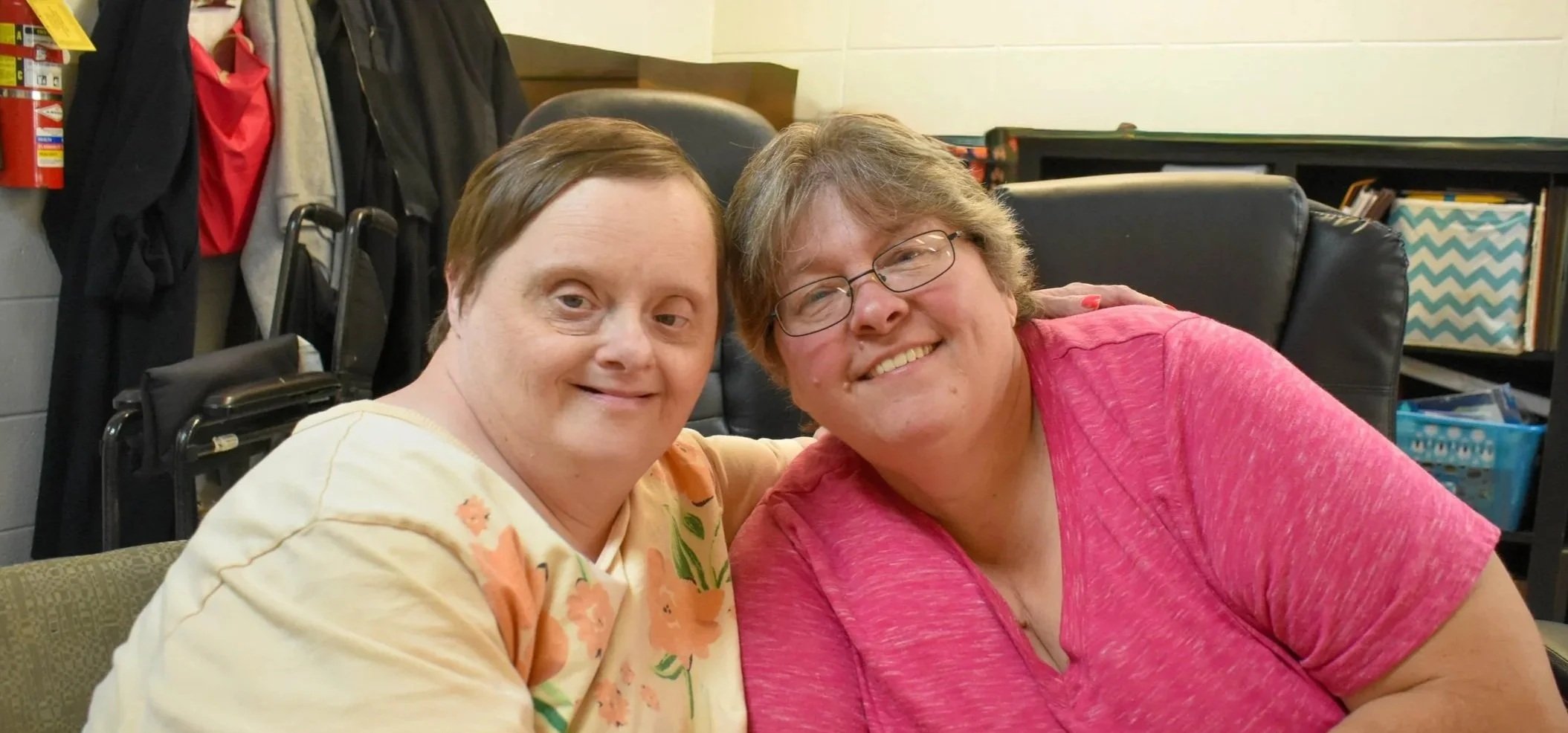 Two women sitting close together, smiling at the camera, in a room with bookshelves and office chairs in the background.