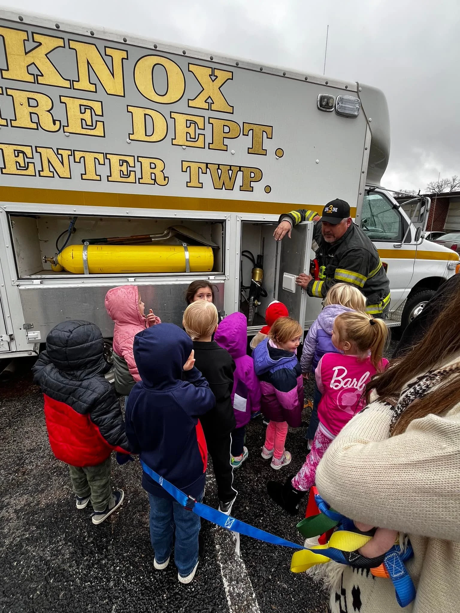 A firefighter giving a demonstration to a group of children next to a fire department truck in a parking lot on a cloudy day.