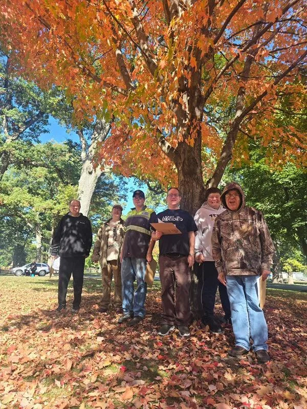 Group of six people standing outdoors under a large autumn tree with orange and green leaves, with fallen leaves on the ground, on a sunny day.