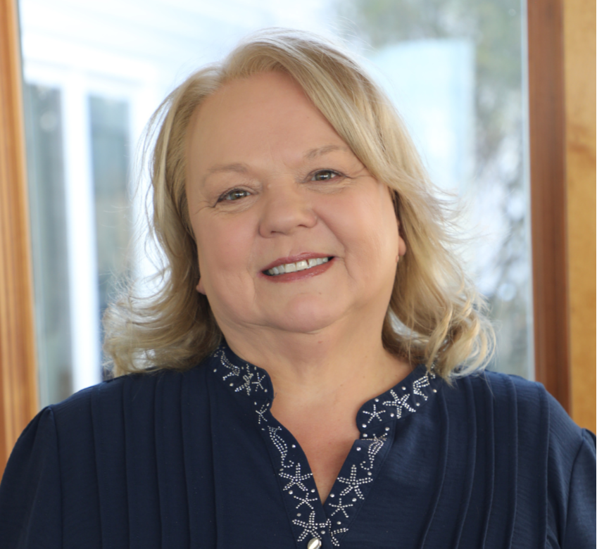 A smiling middle-aged woman with blonde hair, wearing a navy blue top with white decorative stitching, standing indoors near a window with a blurred background of trees.