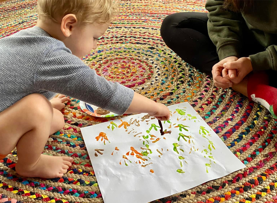 A young child and an adult sitting on a multicolored circular rug, drawing on a piece of white paper with green, orange, and brown markers.