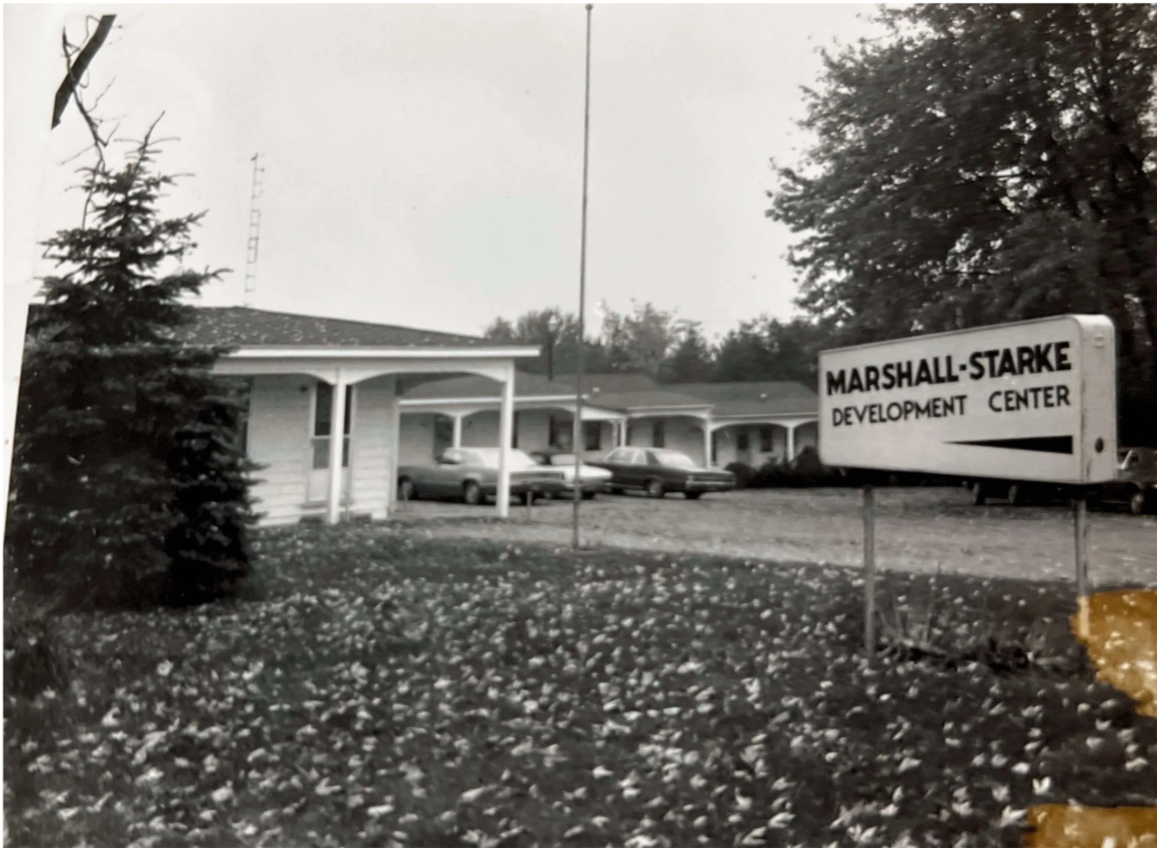 Black and white photograph of the Marshall-Starke Development Center building with trees, cars, and a sign in front that reads 'Marshall-Starke Development Center'.