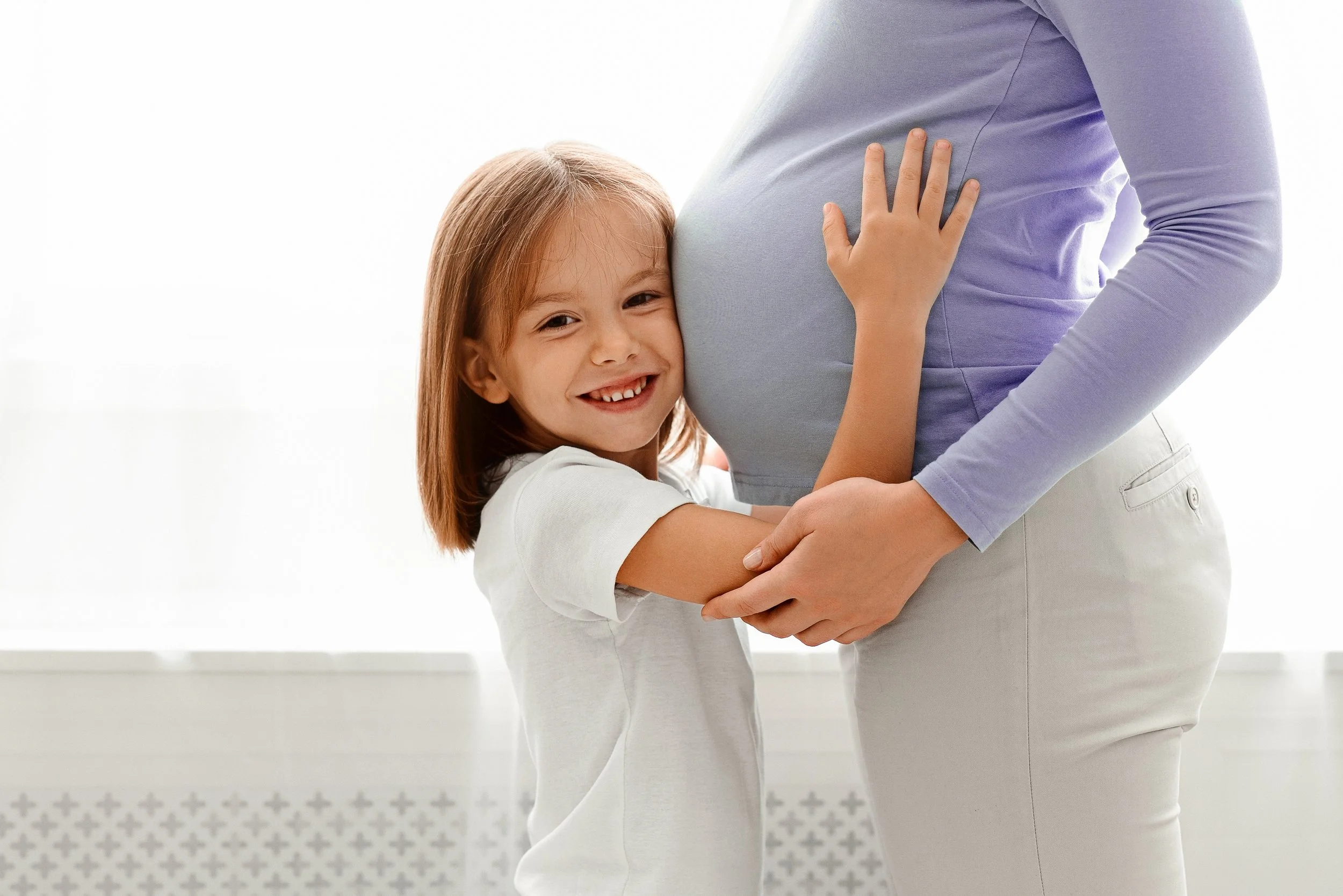 A young girl hugging a pregnant woman, smiling, with her arms around the woman's belly.