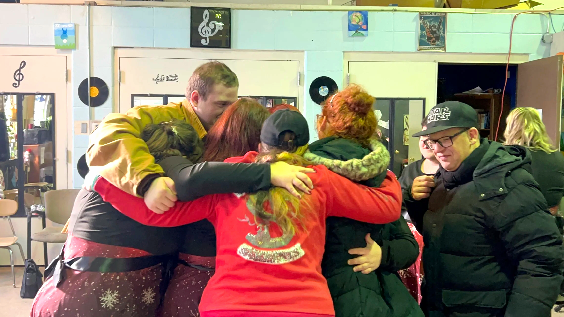 Group of people embracing in a joyful group hug inside a room decorated with music-themed artwork, records, and posters.